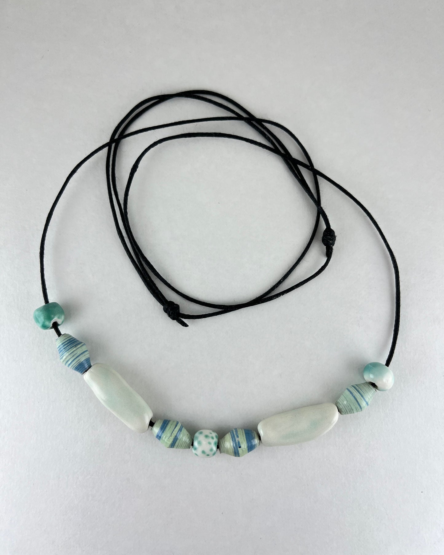 String of porcelain and paper beads on fine cotton thread. 4 paper beads with subtle stripes in light blue and pale green. 3 round porcelain beads in white and minty green, and two chunky pale green porcelain tube beads