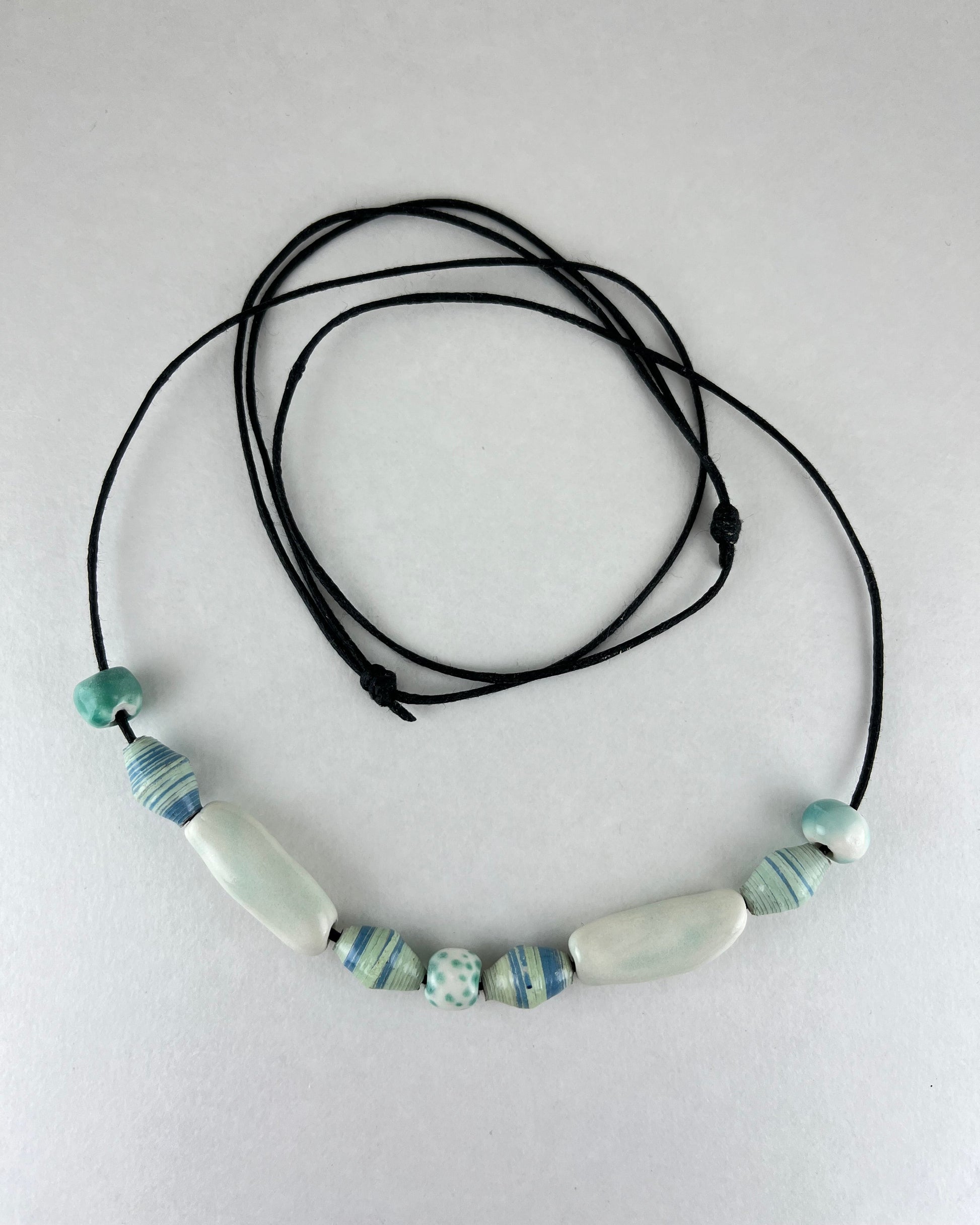 String of porcelain and paper beads on fine cotton thread. 4 paper beads with subtle stripes in light blue and pale green. 3 round porcelain beads in white and minty green, and two chunky pale green porcelain tube beads