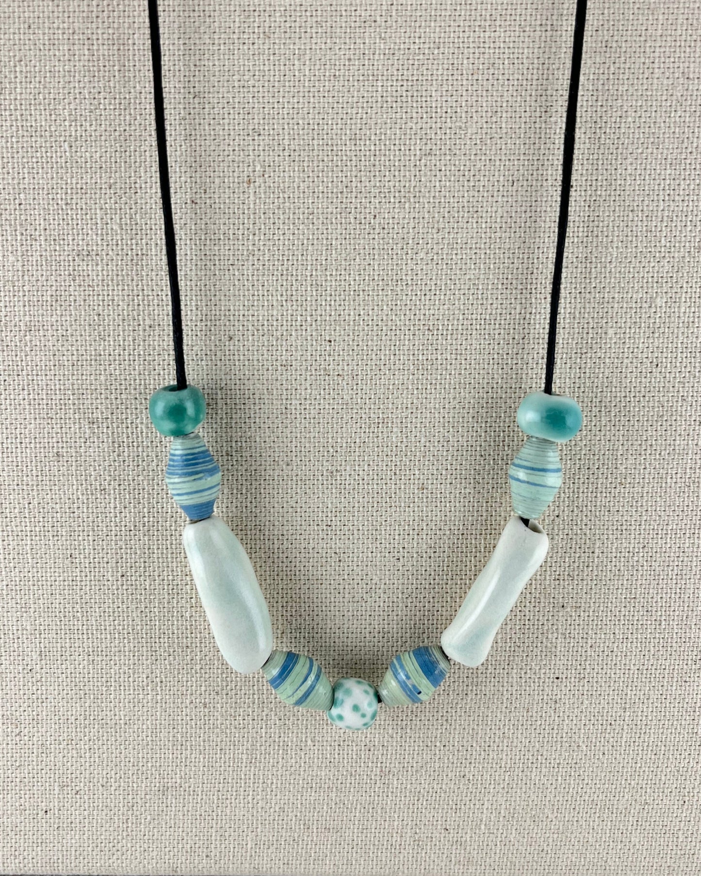 String of porcelain and paper beads on fine cotton thread. 4 paper beads with subtle stripes in light blue and pale green. 3 round porcelain beads in white and minty green, and two chunky pale green porcelain tube beads.