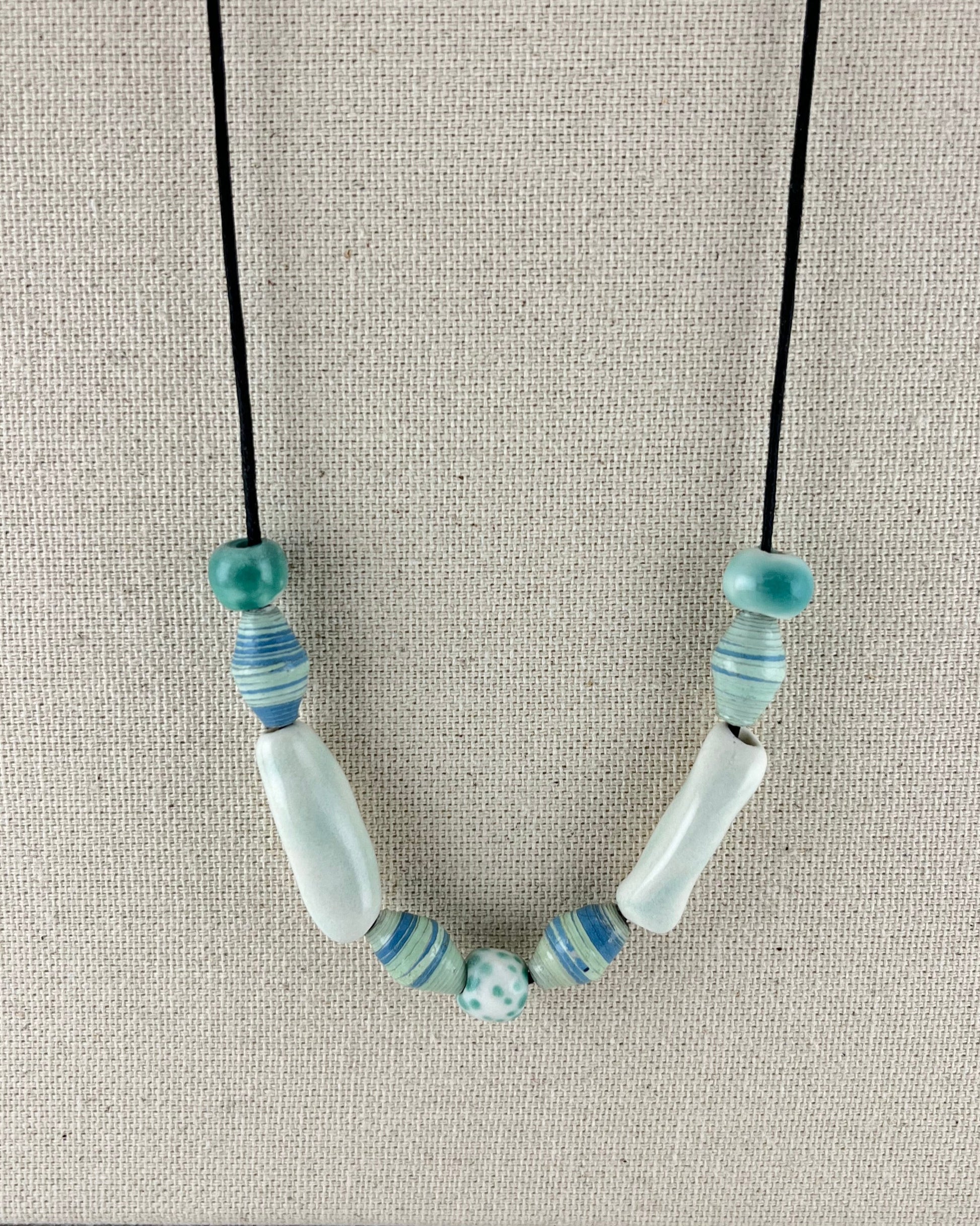 String of porcelain and paper beads on fine cotton thread. 4 paper beads with subtle stripes in light blue and pale green. 3 round porcelain beads in white and minty green, and two chunky pale green porcelain tube beads.