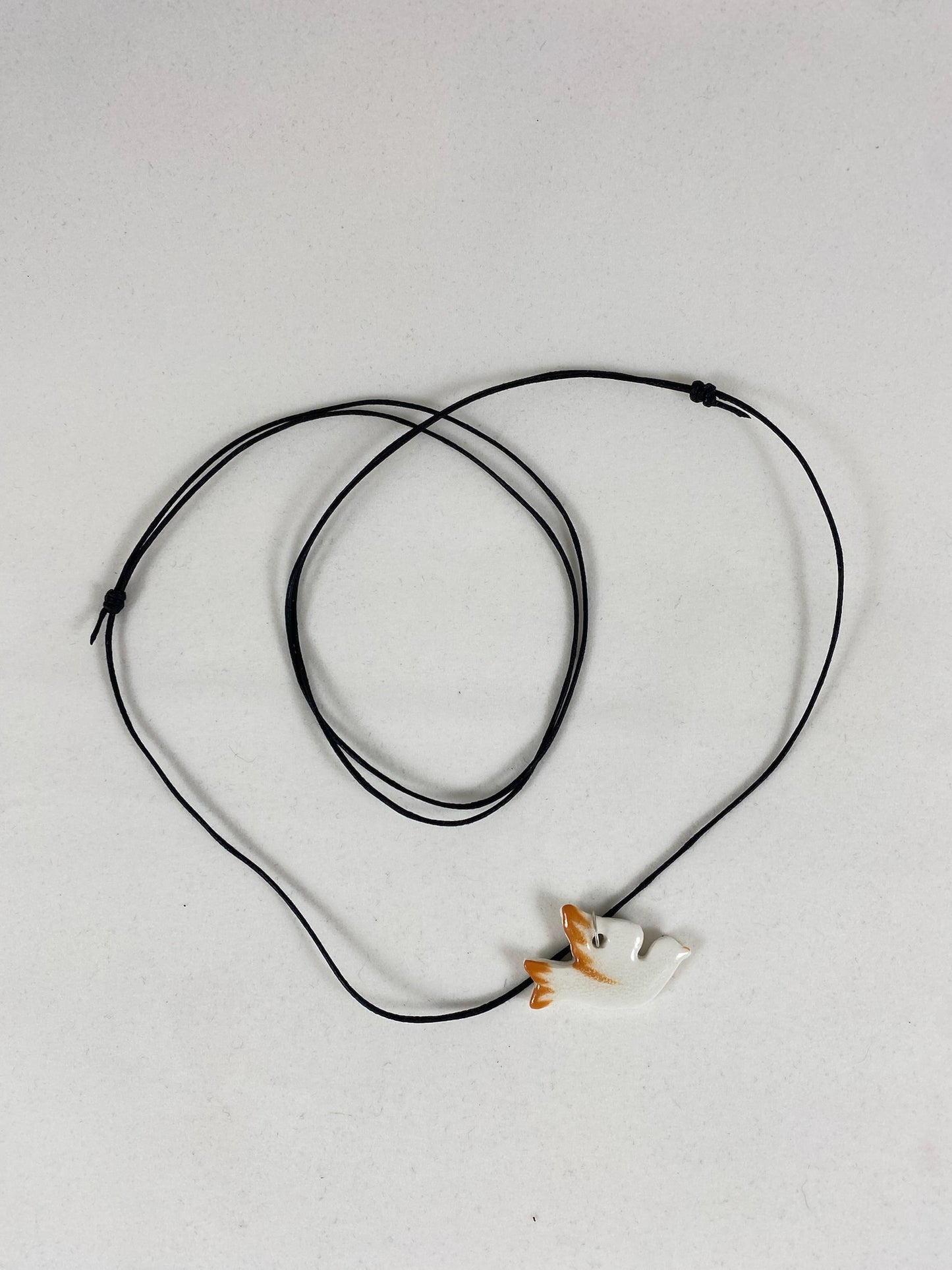 Necklace laid flat on white background. Fine black thread with dove shaped pendant in white with orange wing and tail. Pendant is attached with small sterling silver jump ring.