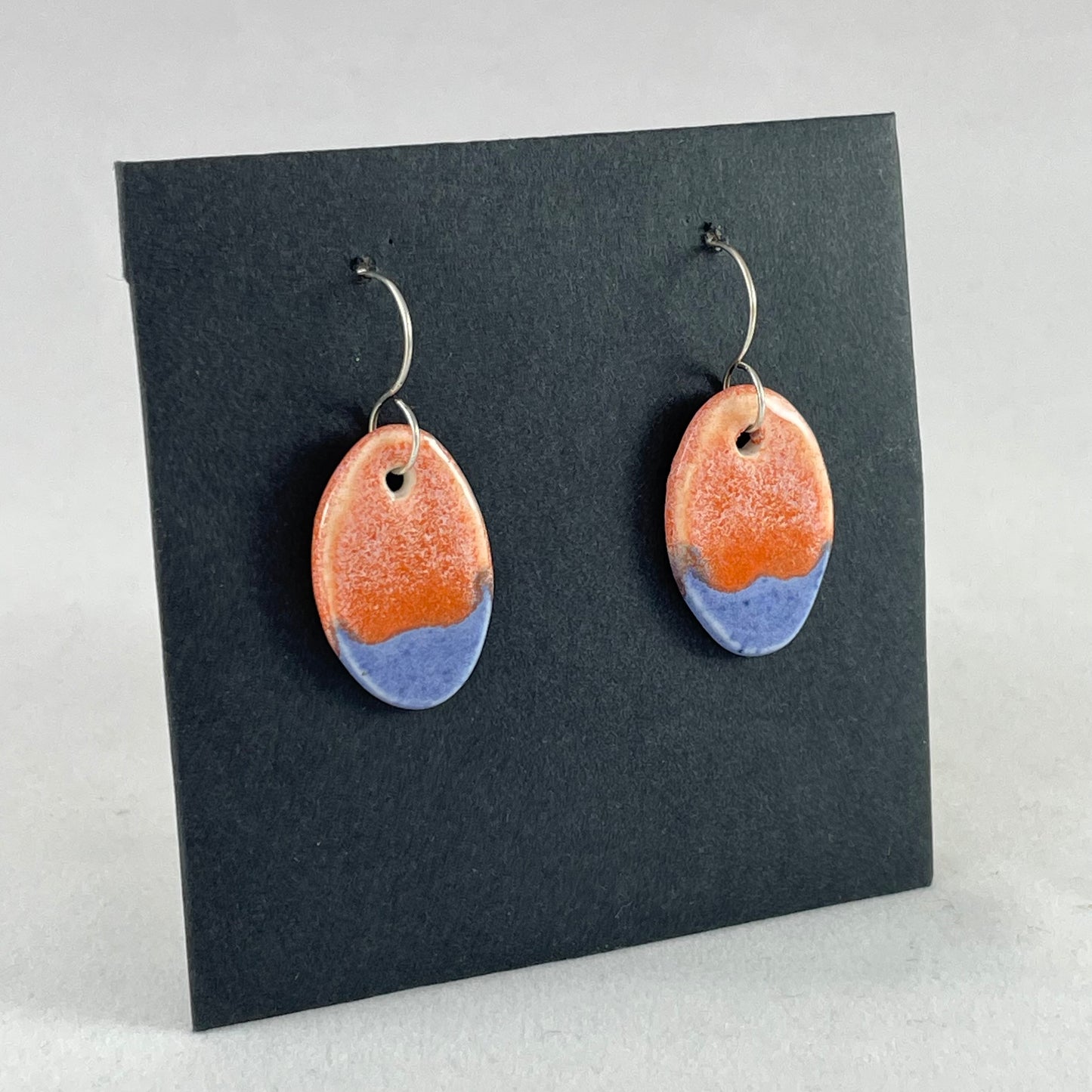 A pair of bright blue and orange oval earrings on a black background. Side view facing right