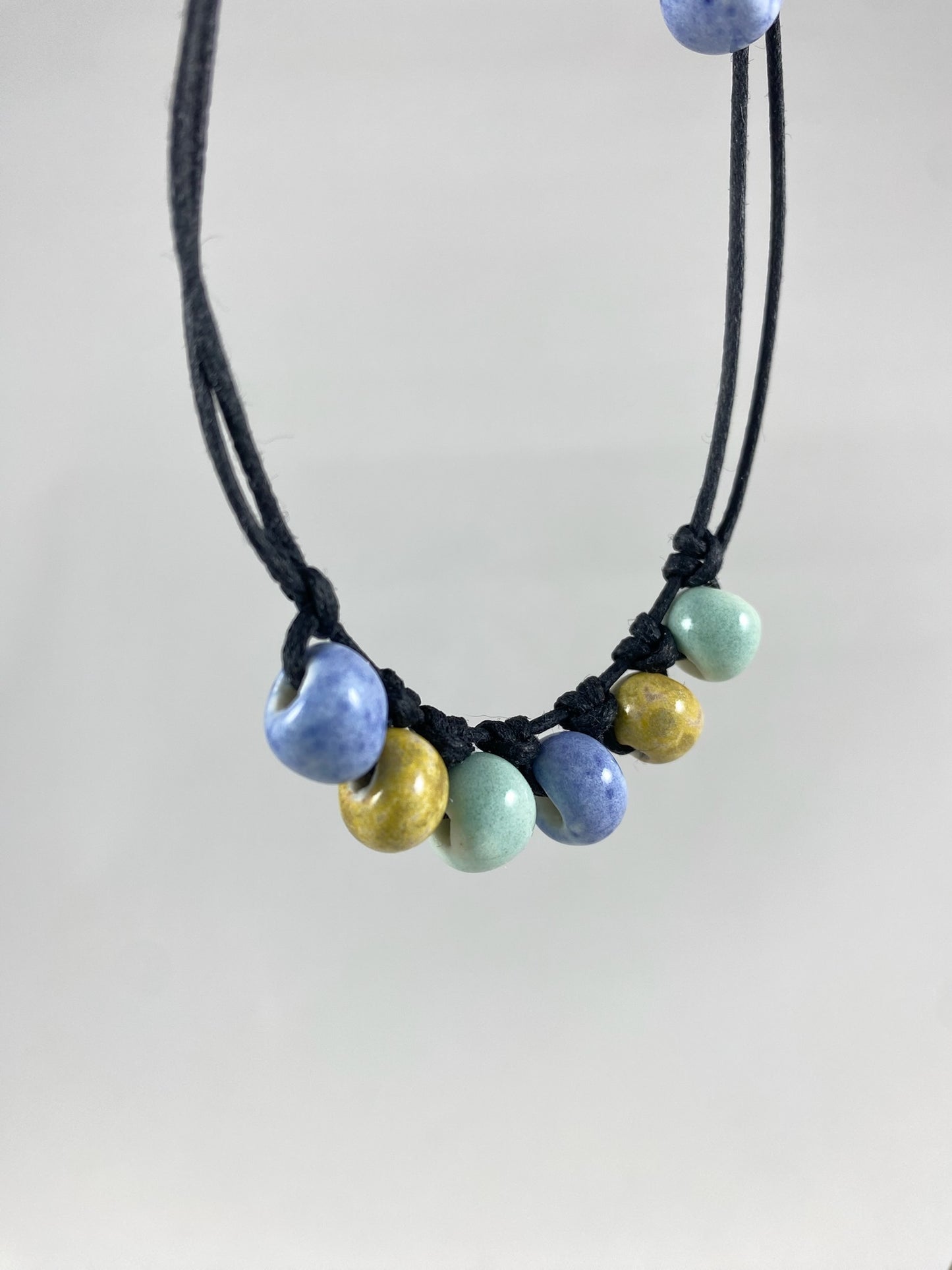 Short necklace hanging against white background. Two strands of fine black string with six porcelain beads attached using simple macrame knots. Two beads are light blue, two are gold and two are light mint green. They are arranged in an alternating order. Photo taken from left hand side.