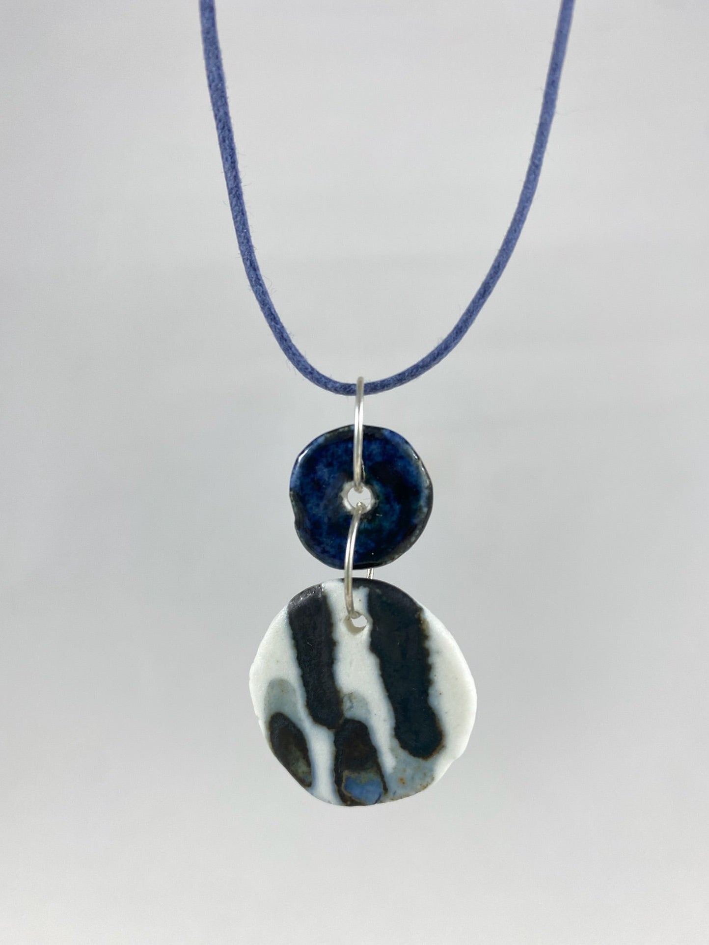Necklace hanging against white background. Fine navy blue string with two circular porcelain pendants attached with silver jump rings. One pendant is small and navy blue and the larger pendant is white with navy glaze drips that looks like watercolour paint.