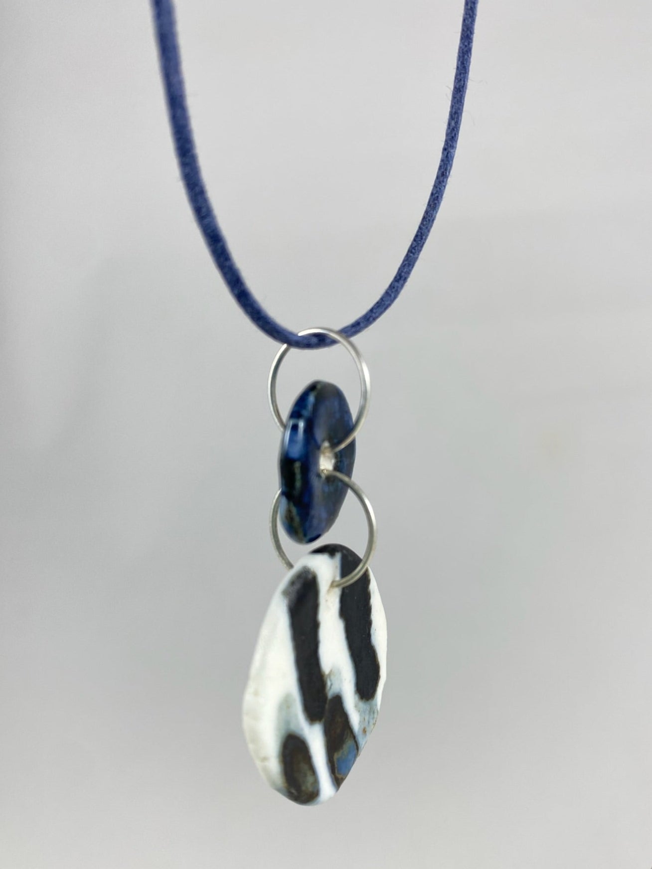 Necklace hanging against white background. Fine navy blue string with two circular porcelain pendants attached with silver jump rings. One pendant is small and navy blue and the larger pendant is white with navy glaze drips that looks like watercolour paint. Side view. Pendants are thin porcelain.
