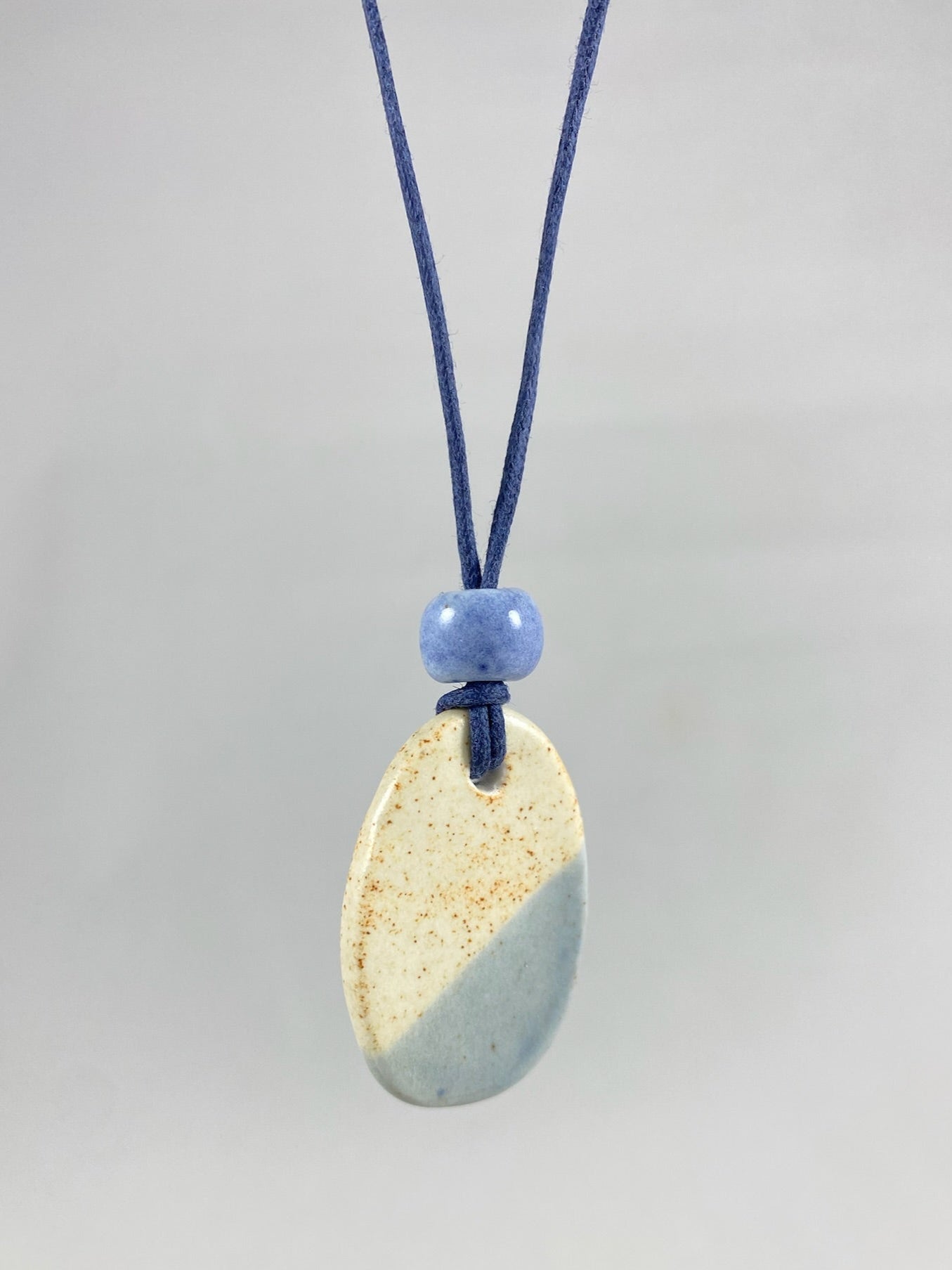 Necklace hanging against white background. Fine navy blue thread with pendant in cream with orange flecks and pale blue. A small pale blue porcelain bead sits above the pendant.