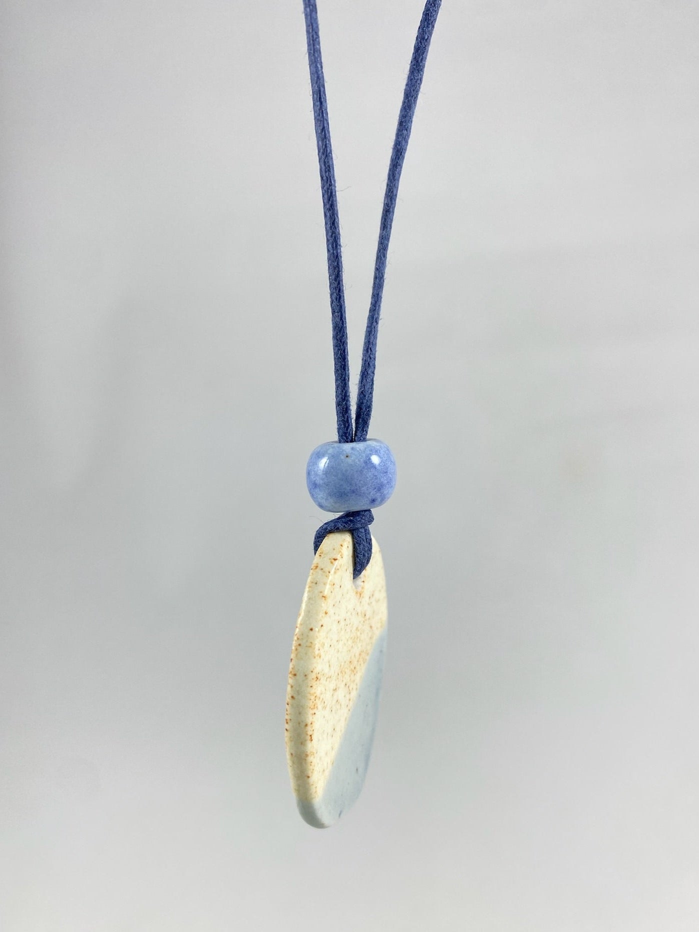 Necklace hanging against white background. Fine navy blue thread with pendant in cream with orange flecks and pale blue. A small pale blue porcelain bead sits above the pendant. Side view. The porcelain pendant is thin.