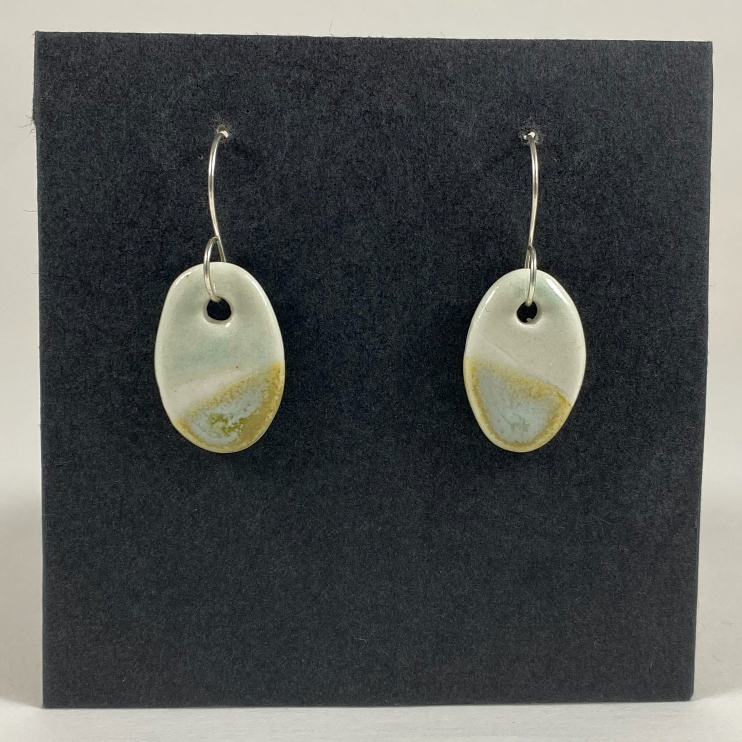 Fine rounded silver hooks with oval shaped pendants in white porcelain. Bottom half dipped asymmetrically in glaze speckled with light green, light brown and pale blue. Top half has a very pale mint green glaze. Photographed on a black background.