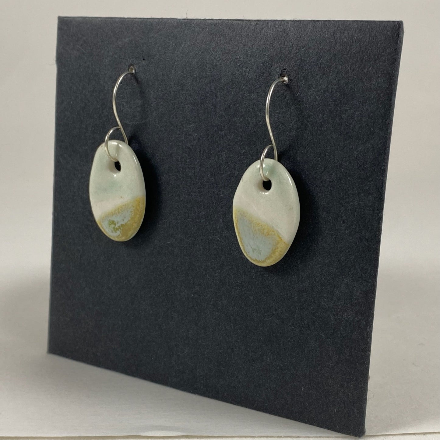 Fine rounded silver hooks with oval shaped pendants in white porcelain. Bottom half dipped asymmetrically in glaze speckled with light green, light brown and pale blue. Photographed on a black background from the right hand side. The porcelain is thin, approximately 2 mm thick.