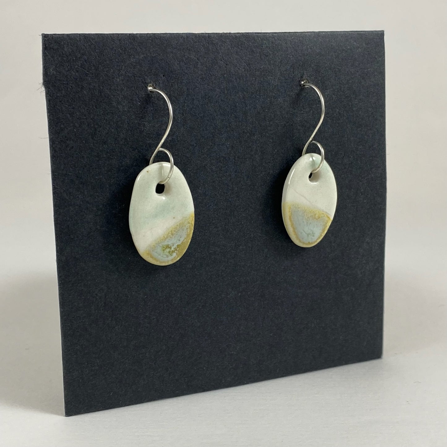 Fine rounded silver hooks with oval shaped pendants in white porcelain. Bottom half dipped asymmetrically in glaze speckled with light green, light brown and pale blue. Photographed on a black background from the left hand side. The porcelain is thin, approximately 2 mm thick.