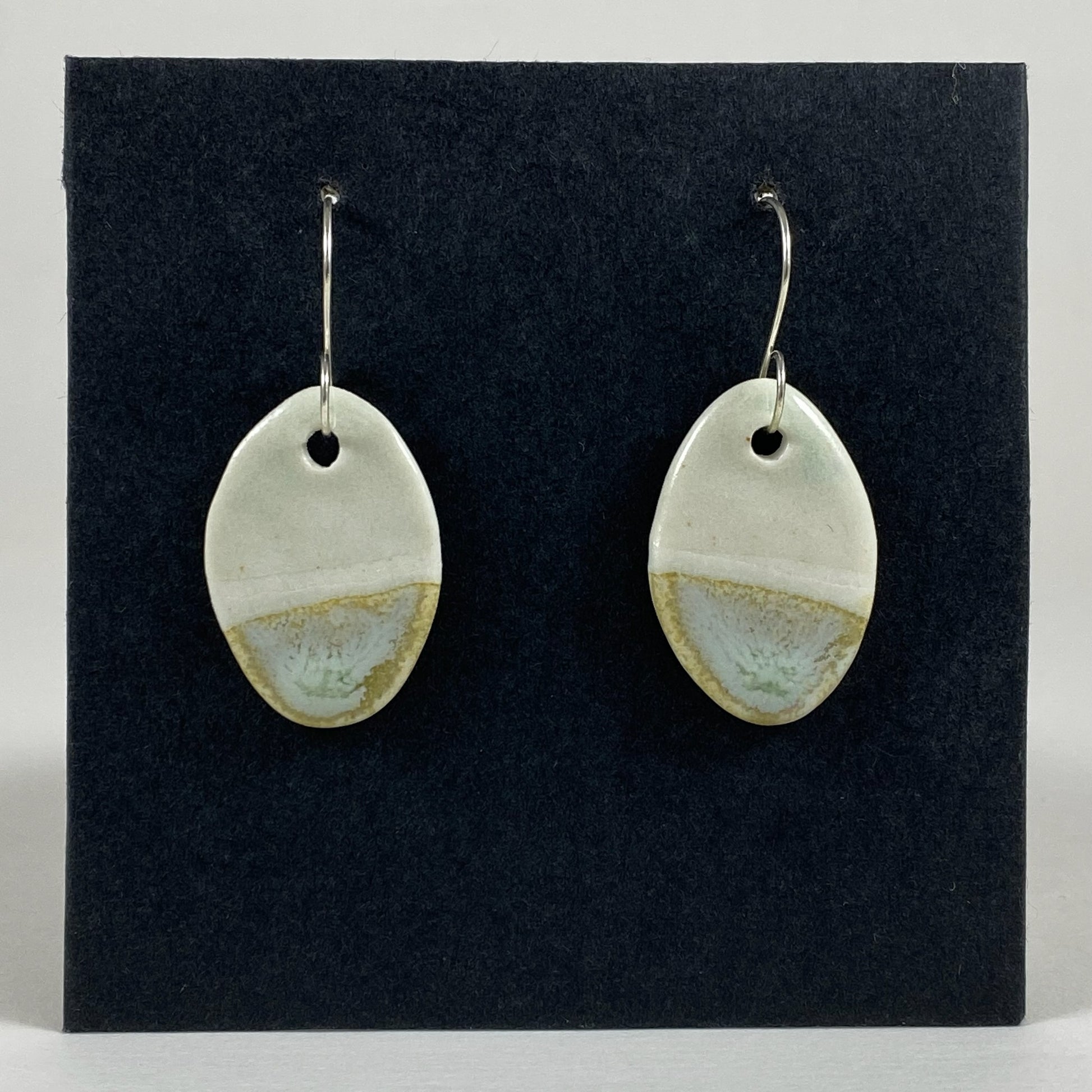 Fine rounded silver hooks with oval shaped pendants in white porcelain. Bottom half dipped asymmetrically in glaze speckled with light green, light brown and pale blue.  Photographed on a black background.