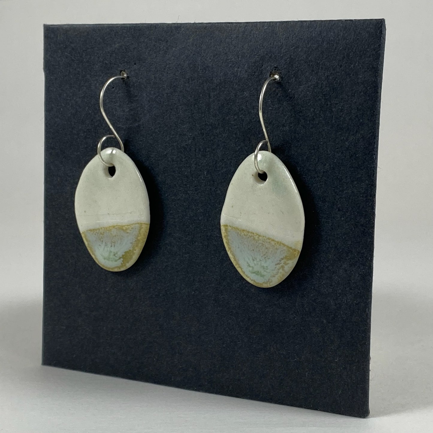 Fine rounded silver hooks with oval shaped pendants in white porcelain. Bottom half dipped in glaze speckled with light green, light brown and pale blue. Photographed on a black background from the right hand side. The porcelain is thin, approximately 3 to 4 mm thick.