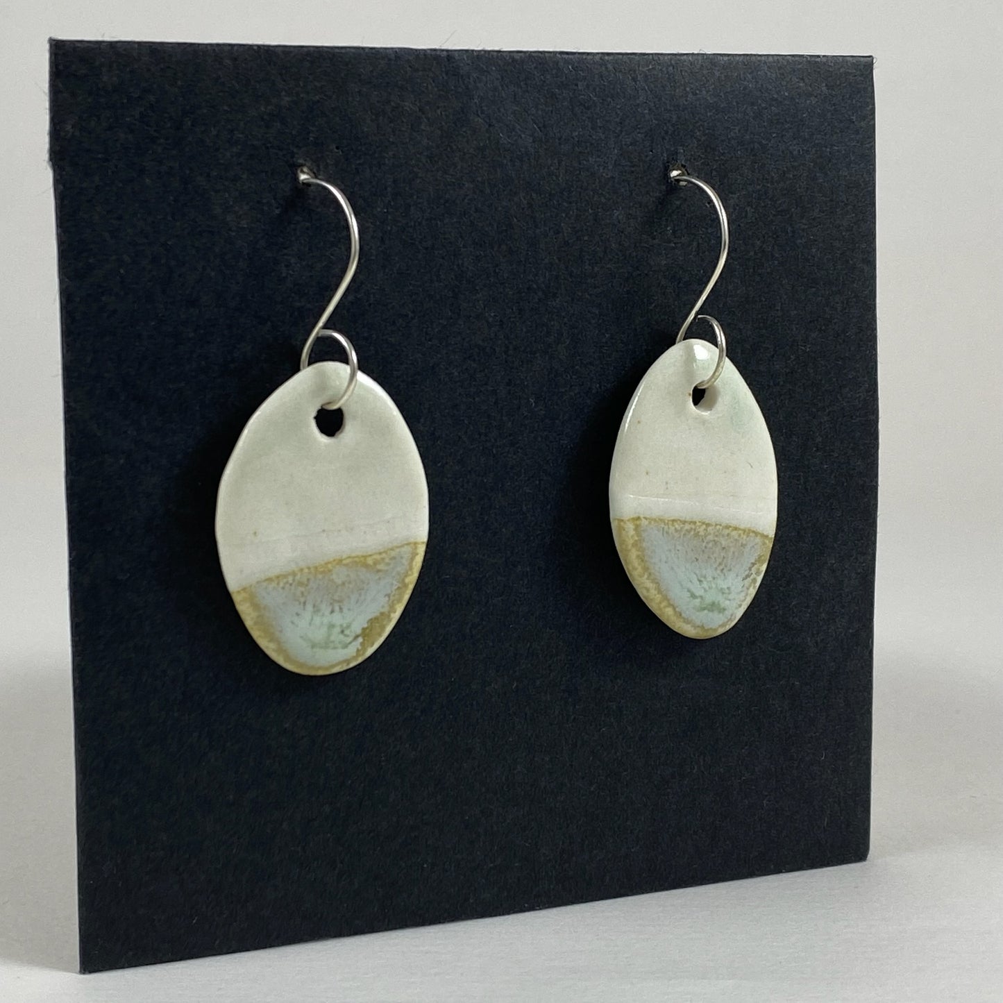 Fine rounded silver hooks with oval shaped pendants in white porcelain. Bottom half dipped in glaze speckled with light green, light brown and pale blue. Photographed on a black background from the left hand side. The porcelain is thin, approximately 3 to 4 mm thick.