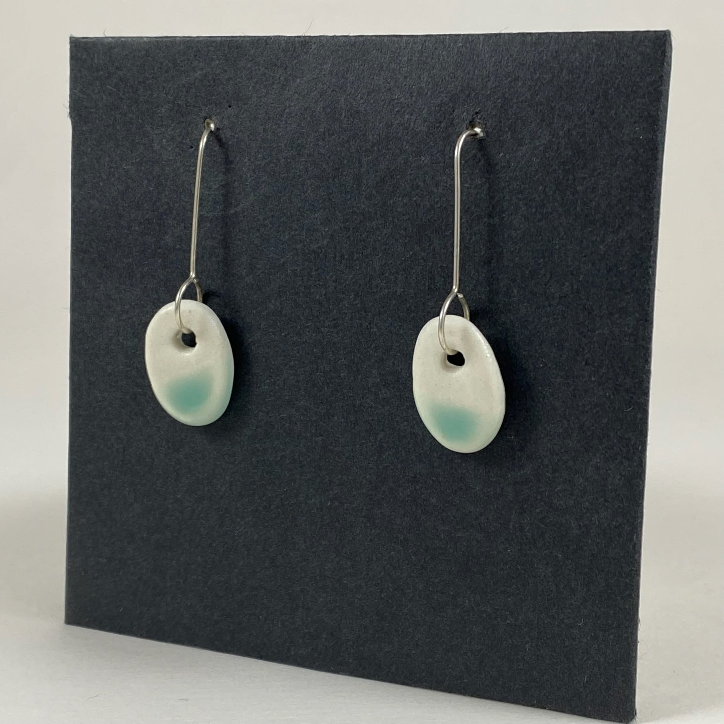 Fine silver hooks with oval shaped pendants in white porcelain. Bottom half dipped in mint green shiny glaze, which fades to white at the edges for a soft look. Photographed on a black background from the side. The porcelain is thin, approximately 2 mm thick.