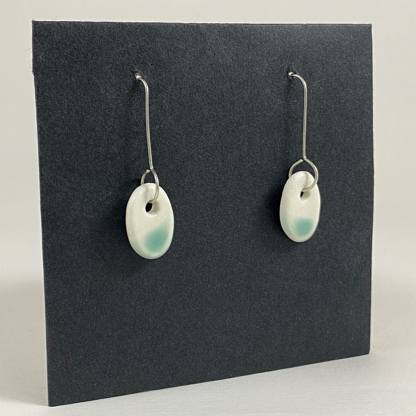 Fine silver hooks with oval shaped pendants in white porcelain. Bottom half dipped in mint green shiny glaze, which fades to white at the edges for a soft look. Photographed on a black background from the left hand side. The porcelain is thin, approximately 2 mm thick.