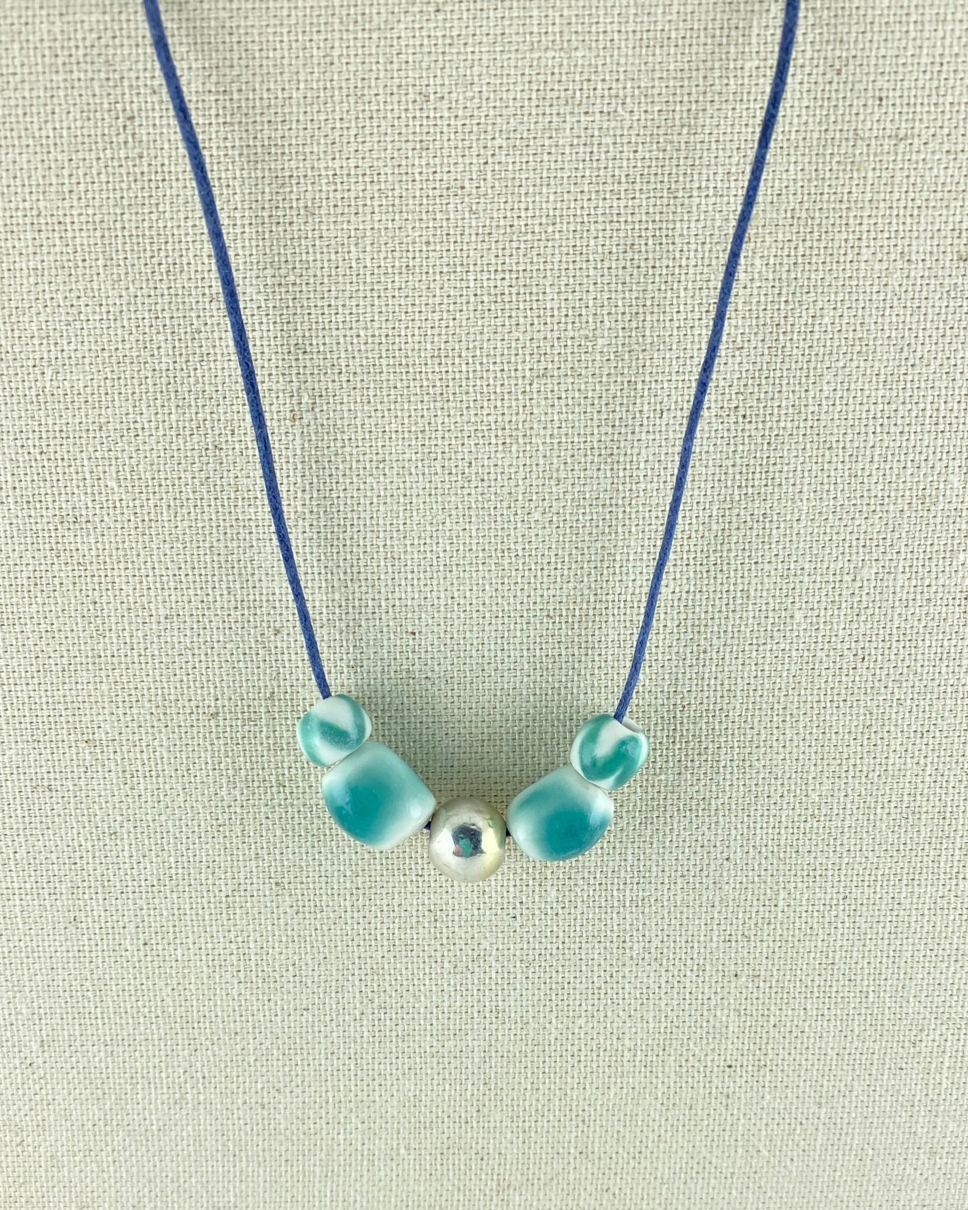 Two small round turquoise beads, two medium sized turquoise beads and a shiny silver bead in the middle. Thread on navy blue thread and photographed hanging against a cream linen background