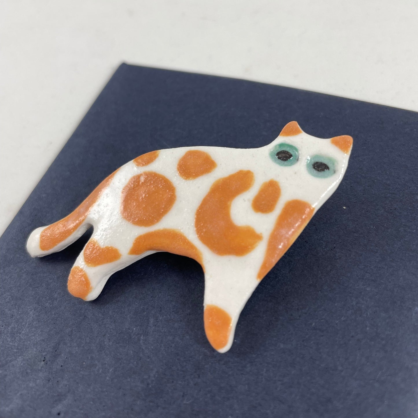White porcelain cat with irregular orange spots and patches. Large green eyes. Photographed flat on a black background from right hand side. Made of thin porcleain.