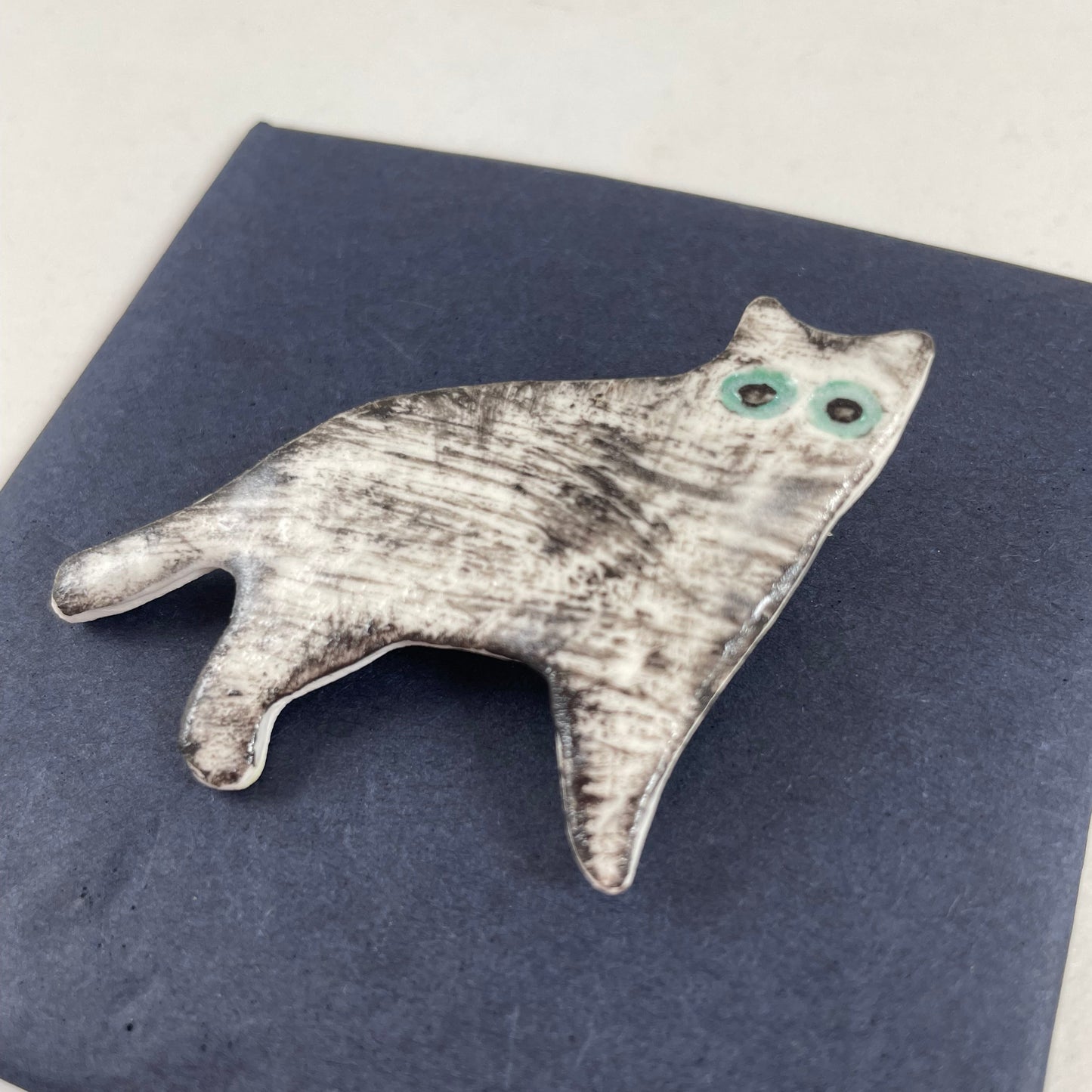 White cat brooch with fine black brush strokes. Effect is grey from a distance or black and white close up. Large green eyes. Photographed on black background.