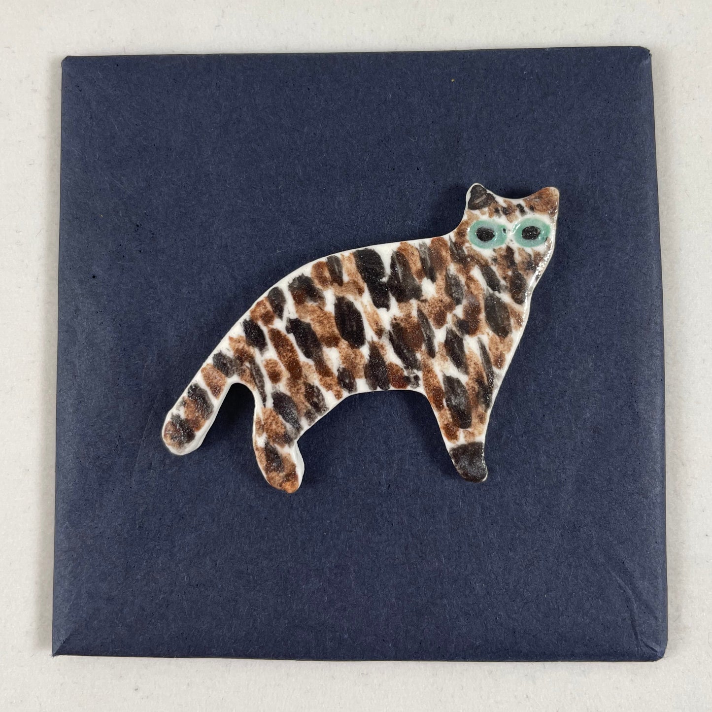 Cat brooch in standing position. Brown and black brush strokes on white porcelain. Effect is like a tiger striped cat. Large green eyes. Photographed on black background.