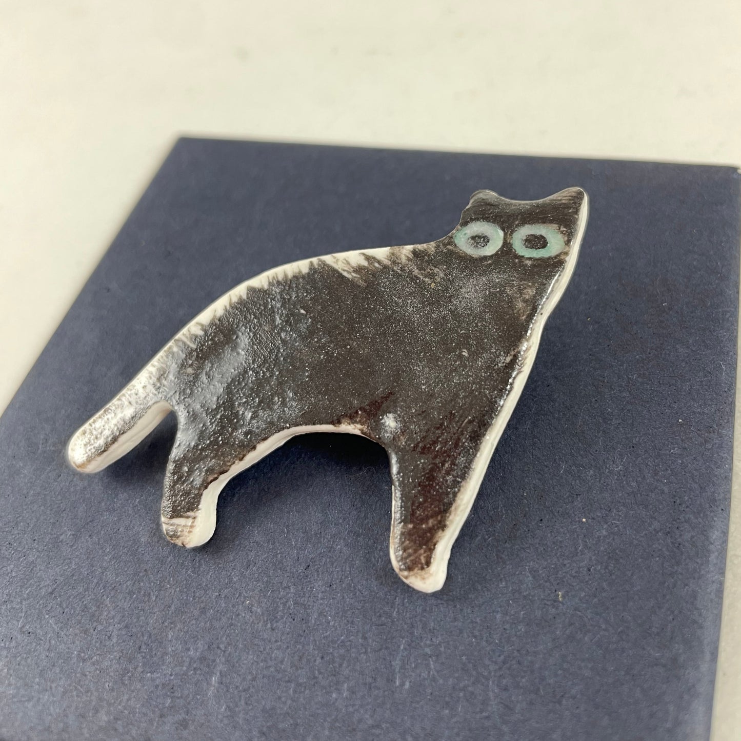 Black porcelain cat. Spiky black brush strokes on its back look like hairs standing on end. Large green eyes. Photographed flat on a black background from right hand side. Made of thin porcelain.