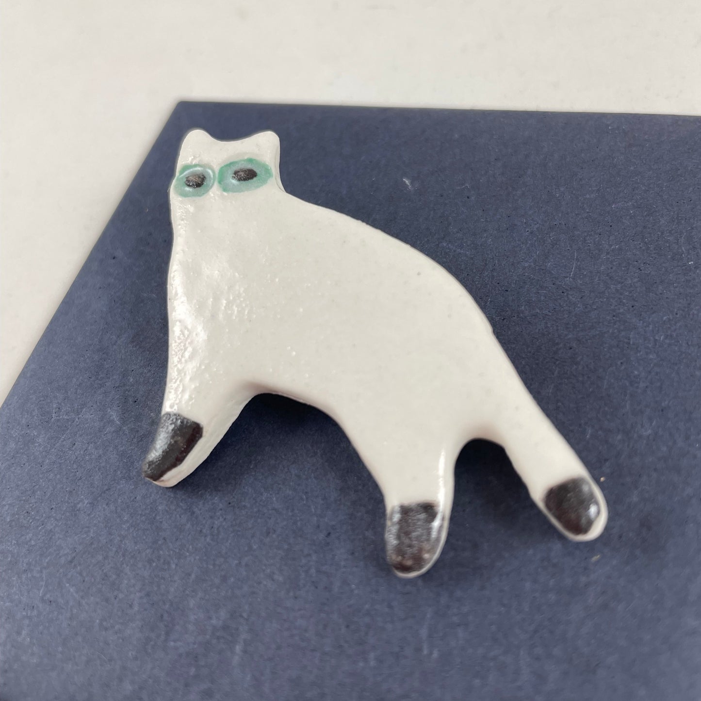 White porcelain cat standing with black feet and tip of tail. Large green eyes. Photographed flat on a black background from right hand side.