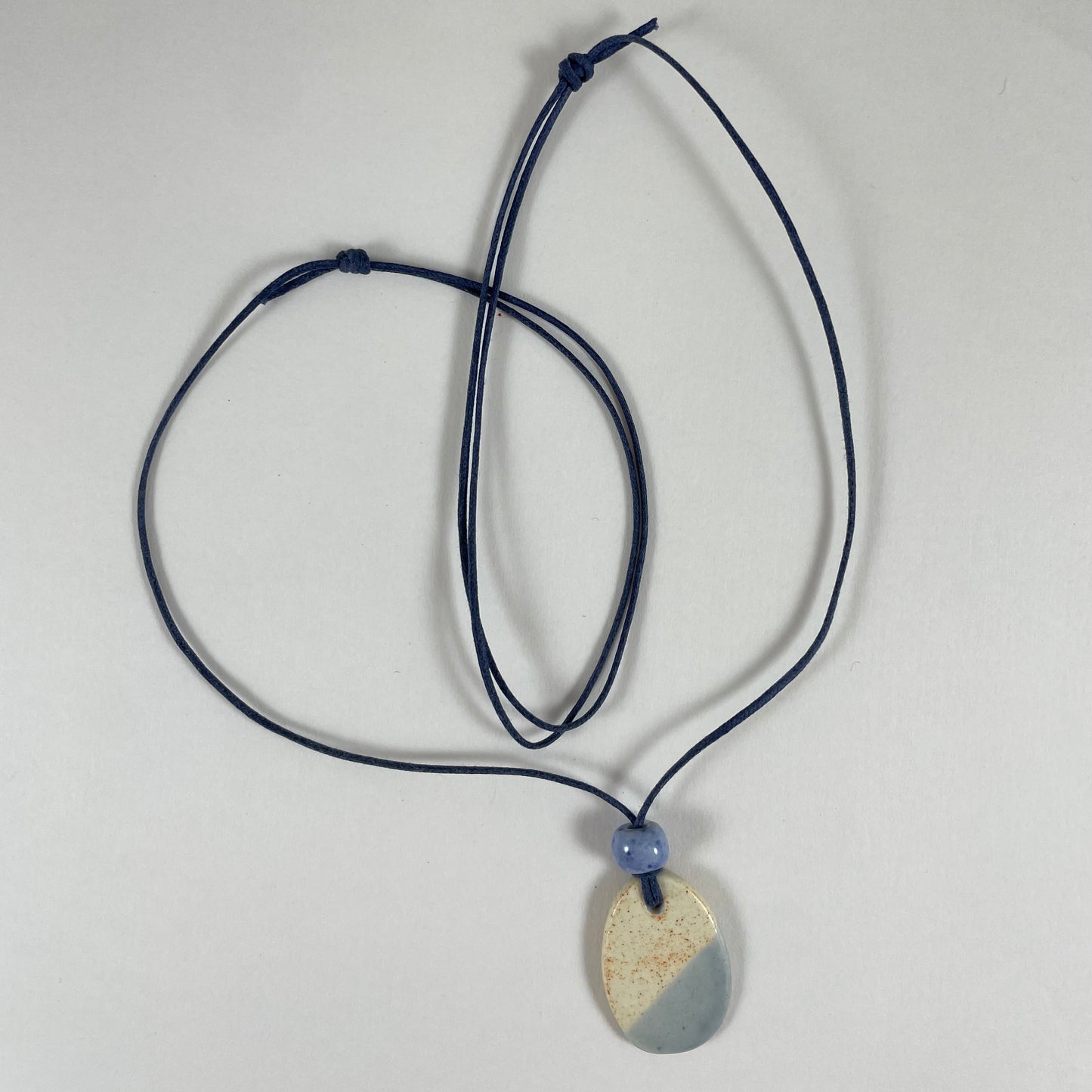 Necklace laid flat on white background. Fine navy blue string with one porcelain pendant and above it a small light blue porcelain bead. The pendant is oval and half cream with tiny orange flecks, and half light blue/lilac.