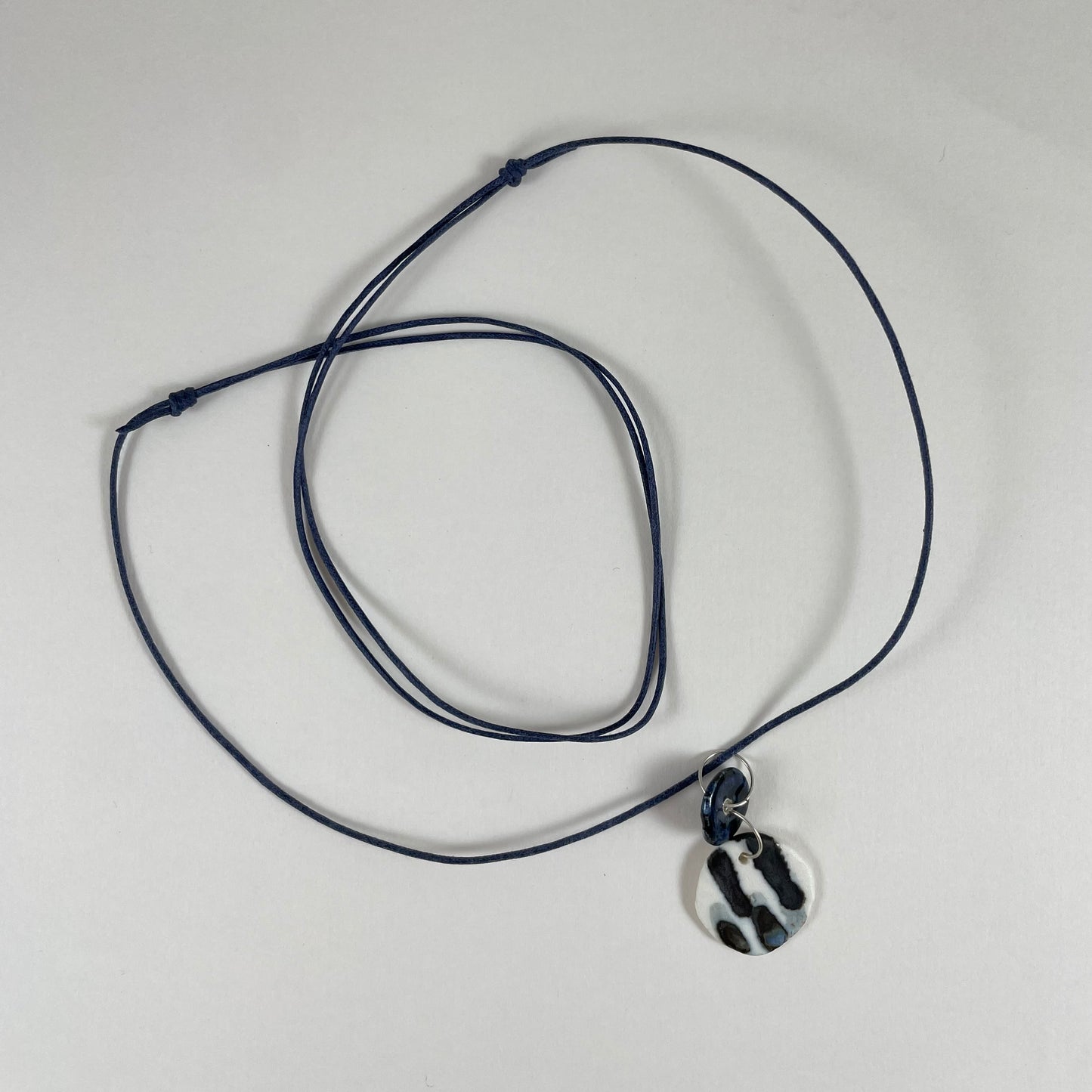 Necklace laid flat on white background. Fine black string with two circular porcelain pendants attached with silver jump rings. One pendant is small and navy blue and the larger pendant is white with navy glaze drips that looks like watercolour paint.