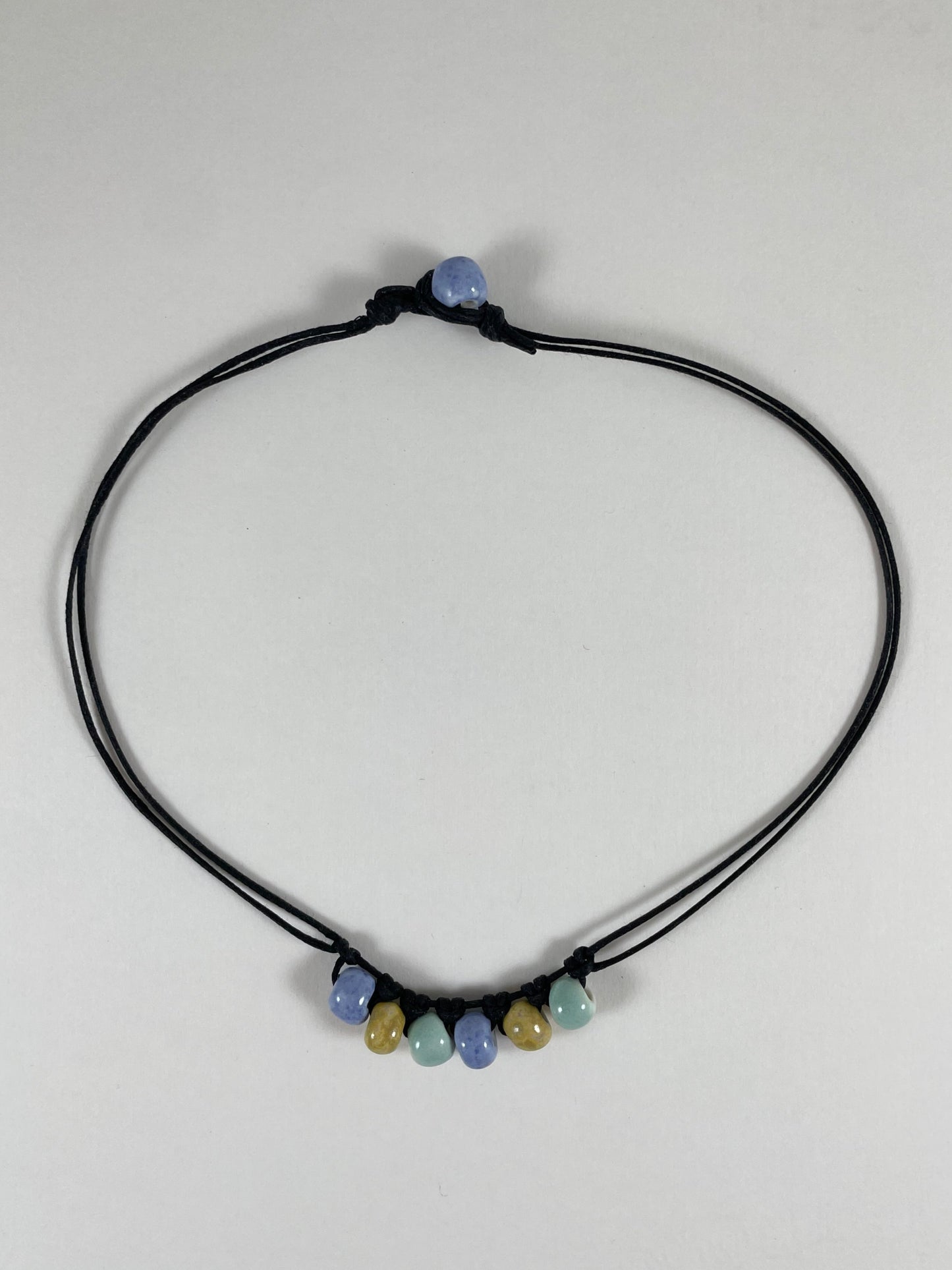 Short necklace laid flat on white background. Two strands of fine black string with six porcelain beads attached using simple macrame knots. Two beads are light blue, two are gold and two are light mint green. They are arranged in an alternating order. One end of necklace has a blue bead attached and the other end has a small loop for the bead to attach to.