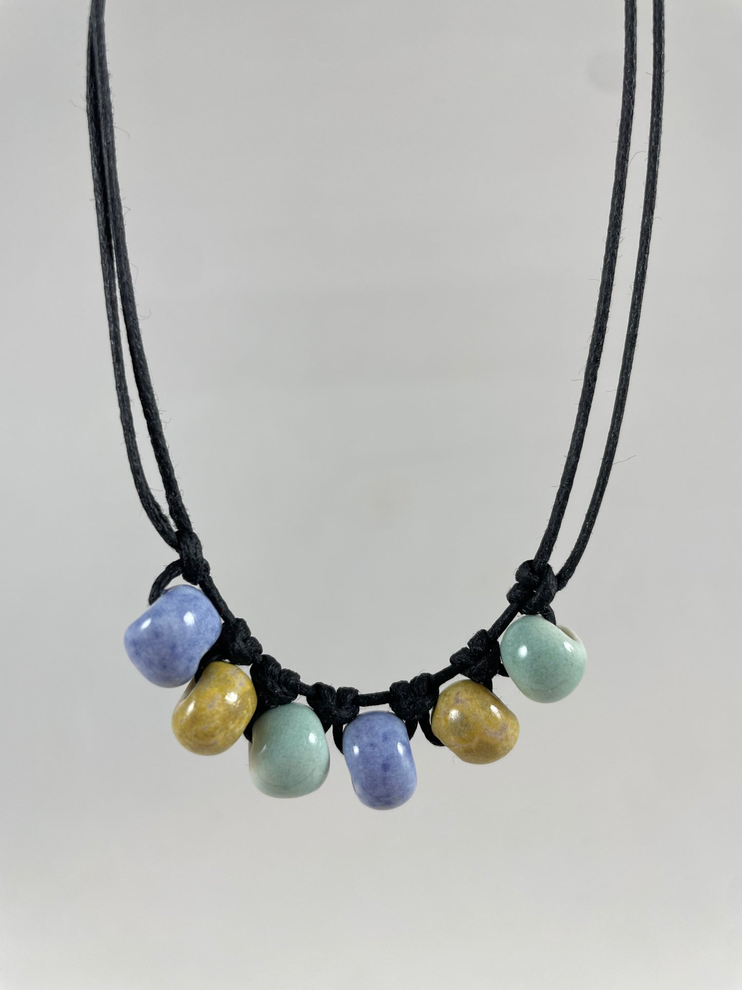 Short necklace hanging against white background. Two strands of fine black string with six porcelain beads attached using simple macrame knots. Two beads are light blue, two are gold and two are light mint green. They are arranged in an alternating order.