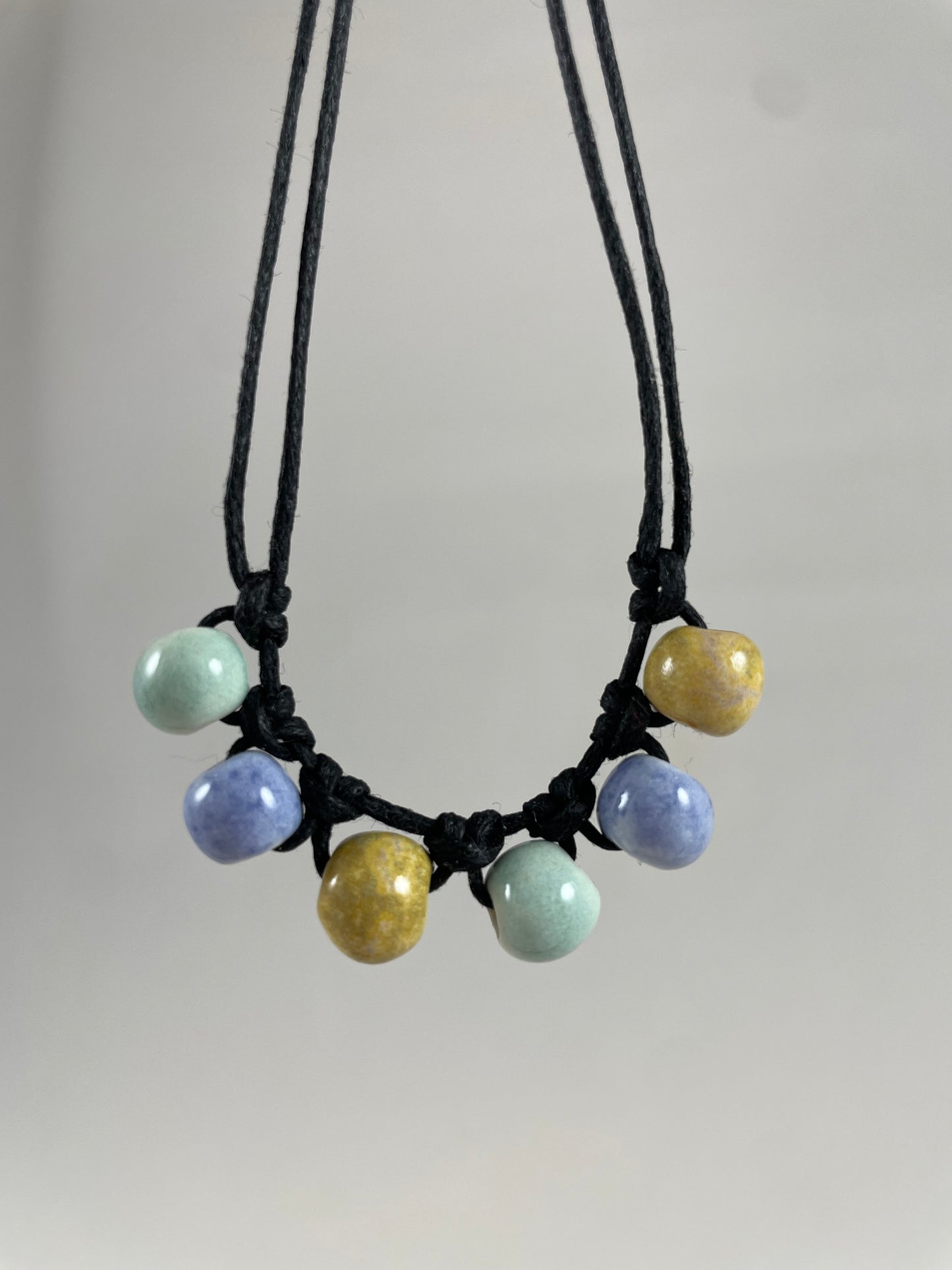 Short necklace hanging against white background. Two strands of fine black string with six porcelain beads attached using simple macrame knots. Two beads are light blue, two are gold and two are light mint green. They are arranged in an alternating order.
