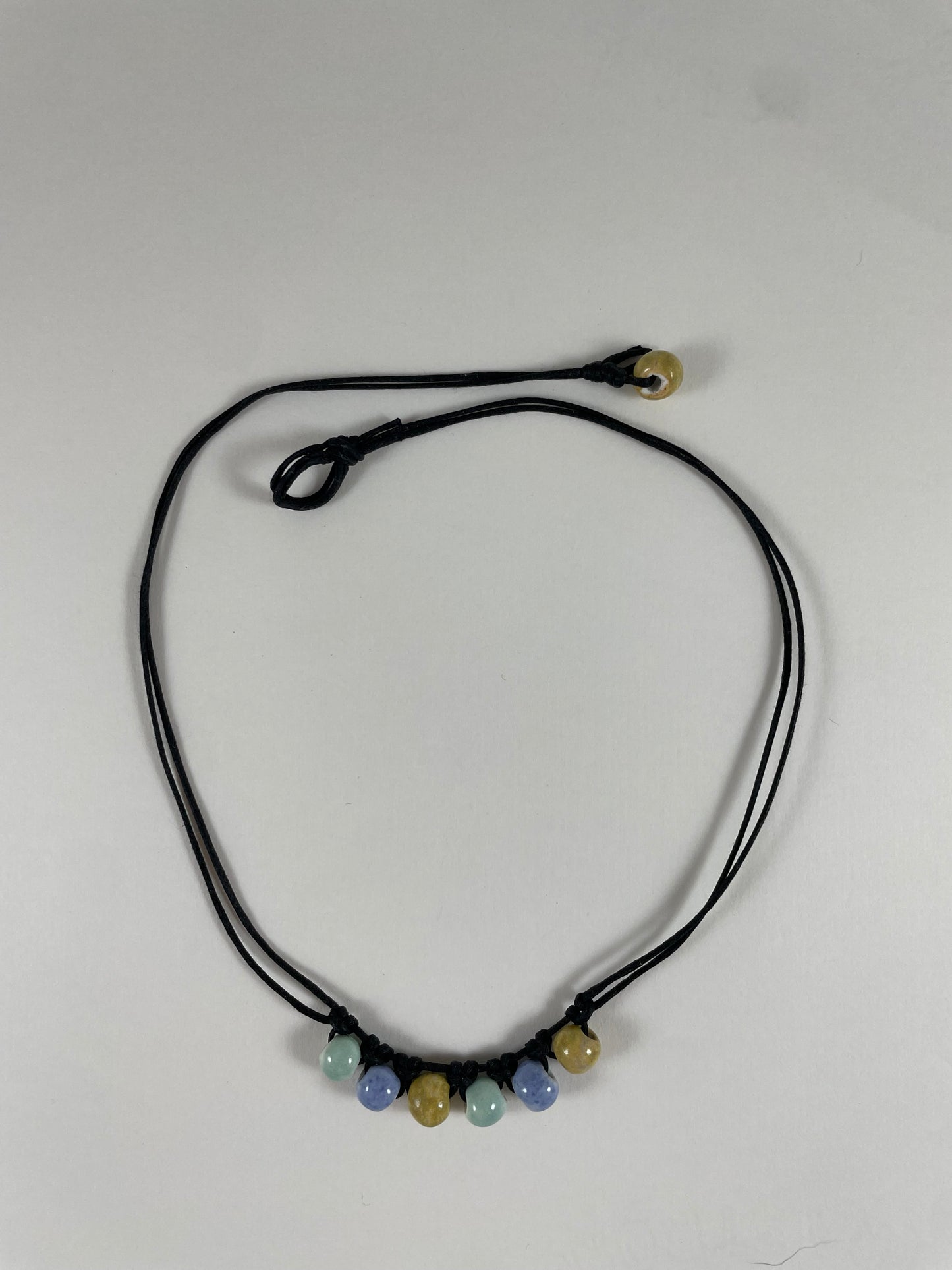 Short necklace laid flat on white background. Two strands of fine black string with six porcelain beads attached using simple macrame knots. Two beads are light blue, two are gold and two are light mint green. They are arranged in an alternating order. One end of necklace has a gold bead attached and the other end has a small loop for the bead to attach to.
