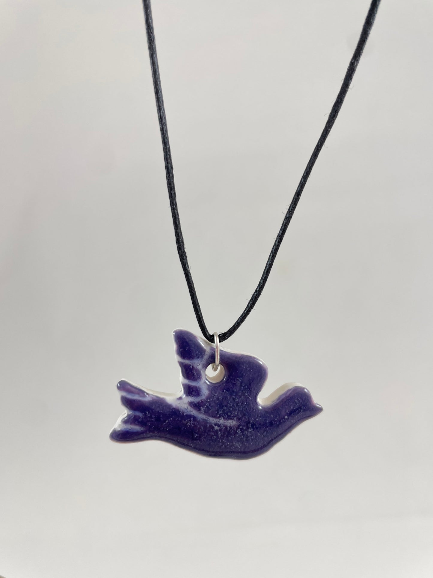 Necklace hanging against white background. Fine black thread with dove shaped pendant in dark lavender purple with fine white etching on wing and tail. Pendant is attached with small sterling silver jump ring.