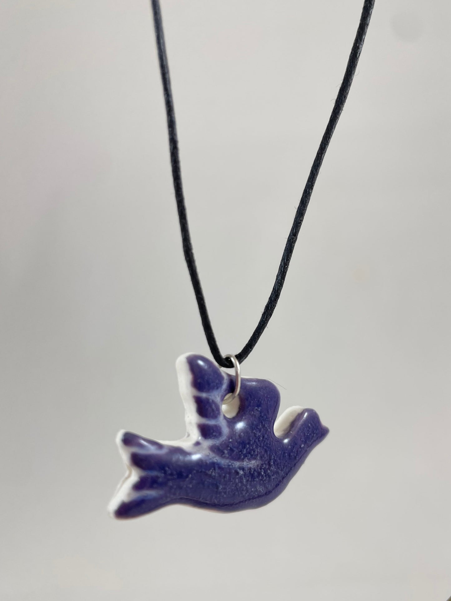 Necklace hanging against white background. Fine black thread with dove shaped pendant in dark lavender purple with fine white etching on wing and tail. Pendant is attached with small sterling silver jump ring. Side view. Pendant is made of thin porcelain.