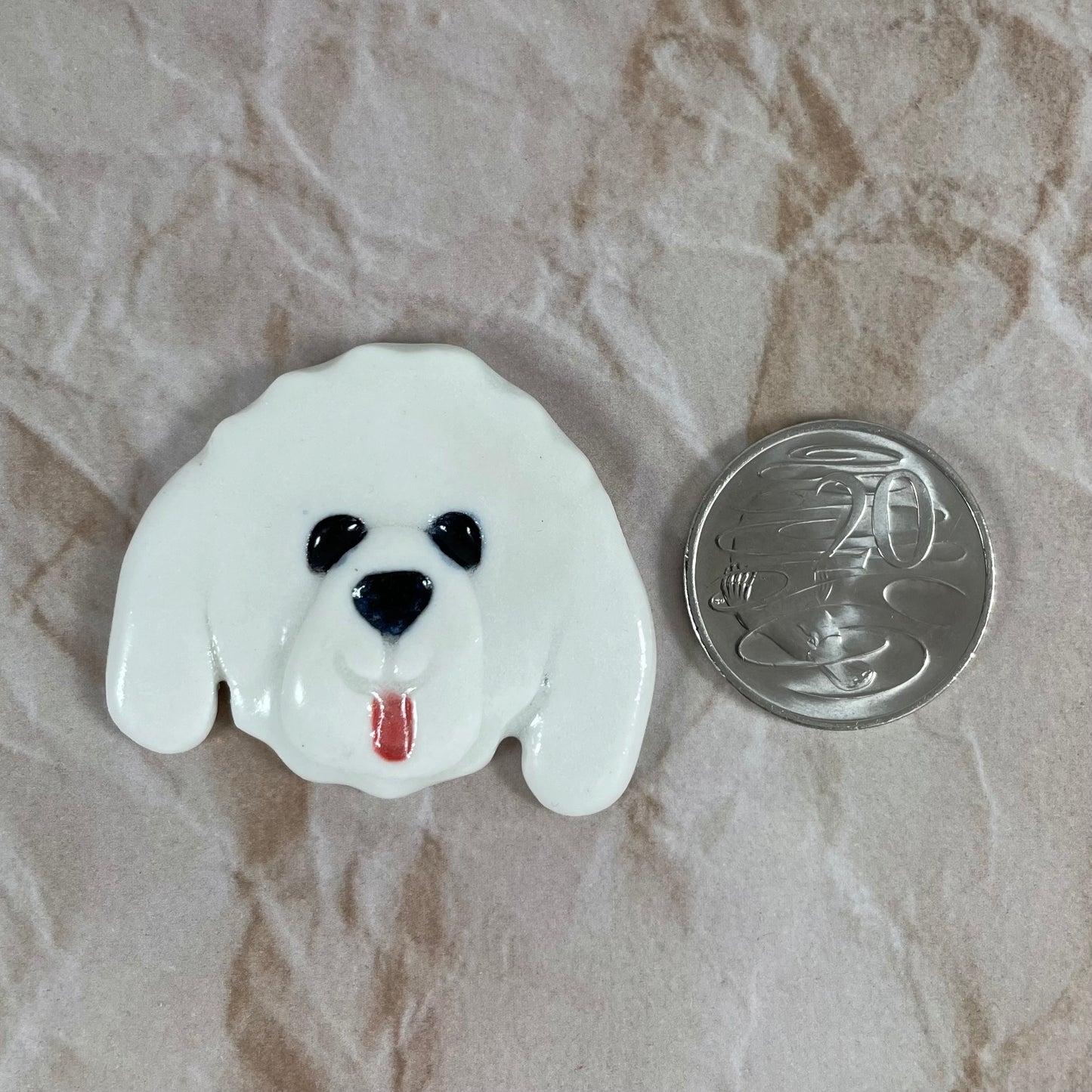 Dog's head in glossy white porcelain with long ears and pink tongue sticking out. Shiny black eyes and nose. Muzzle area is slightly raised to stand out. Photographed next to 20 cent coin, which is smaller than brooch.