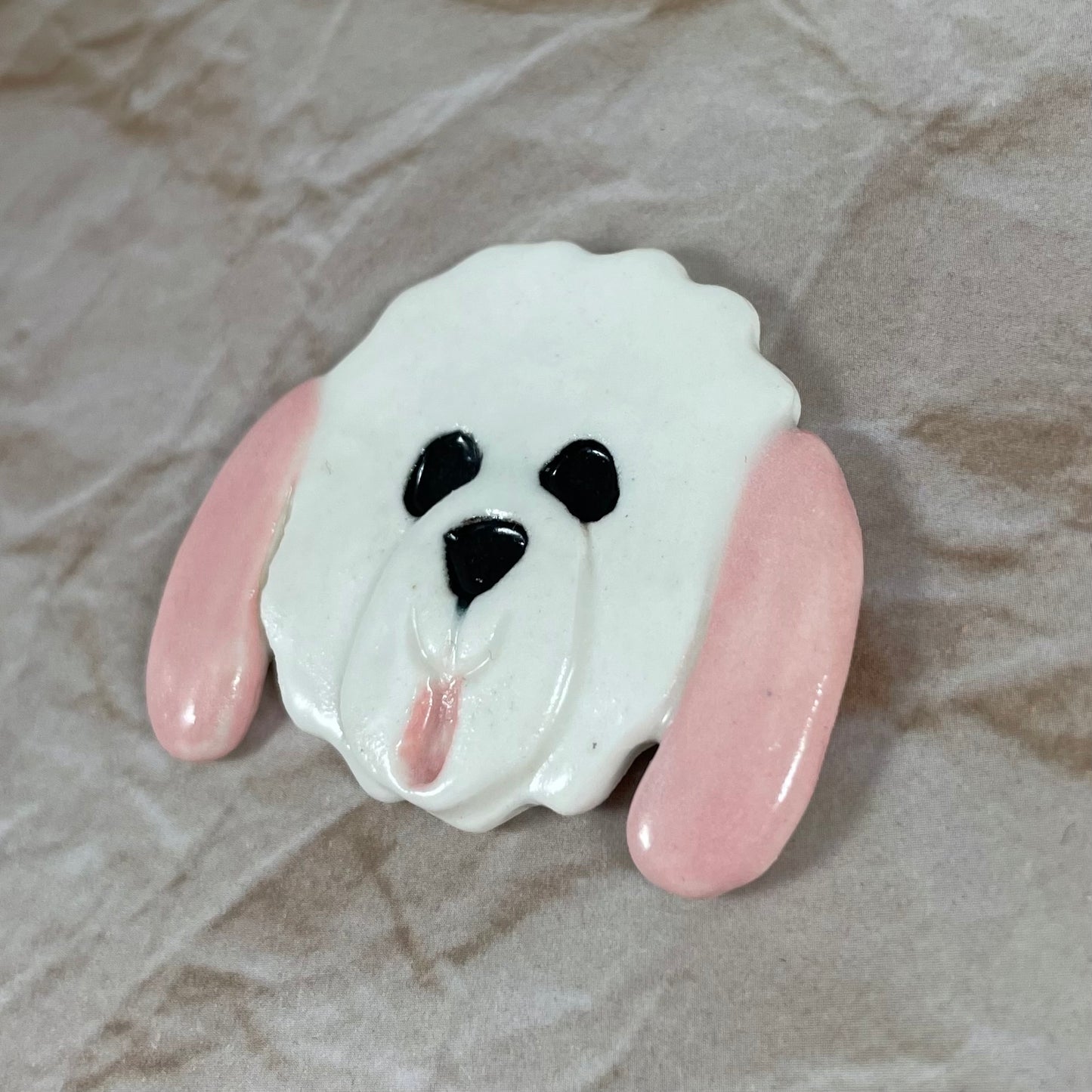 Dog's head in glossy white porcelain with long pale pink ears and pink tongue sticking out. Shiny black eyes and nose. Muzzle area is slightly raised to stand out. Photographed from right hand side. Porcelain is thin