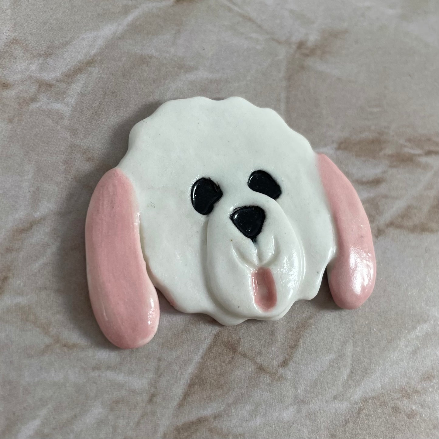 Dog's head in glossy white porcelain with pale pink long ears and rosy pink tongue sticking out. Shiny black eyes and nose. Muzzle area is slightly raised to stand out. Photographed from left hand side. Porcelain is thin