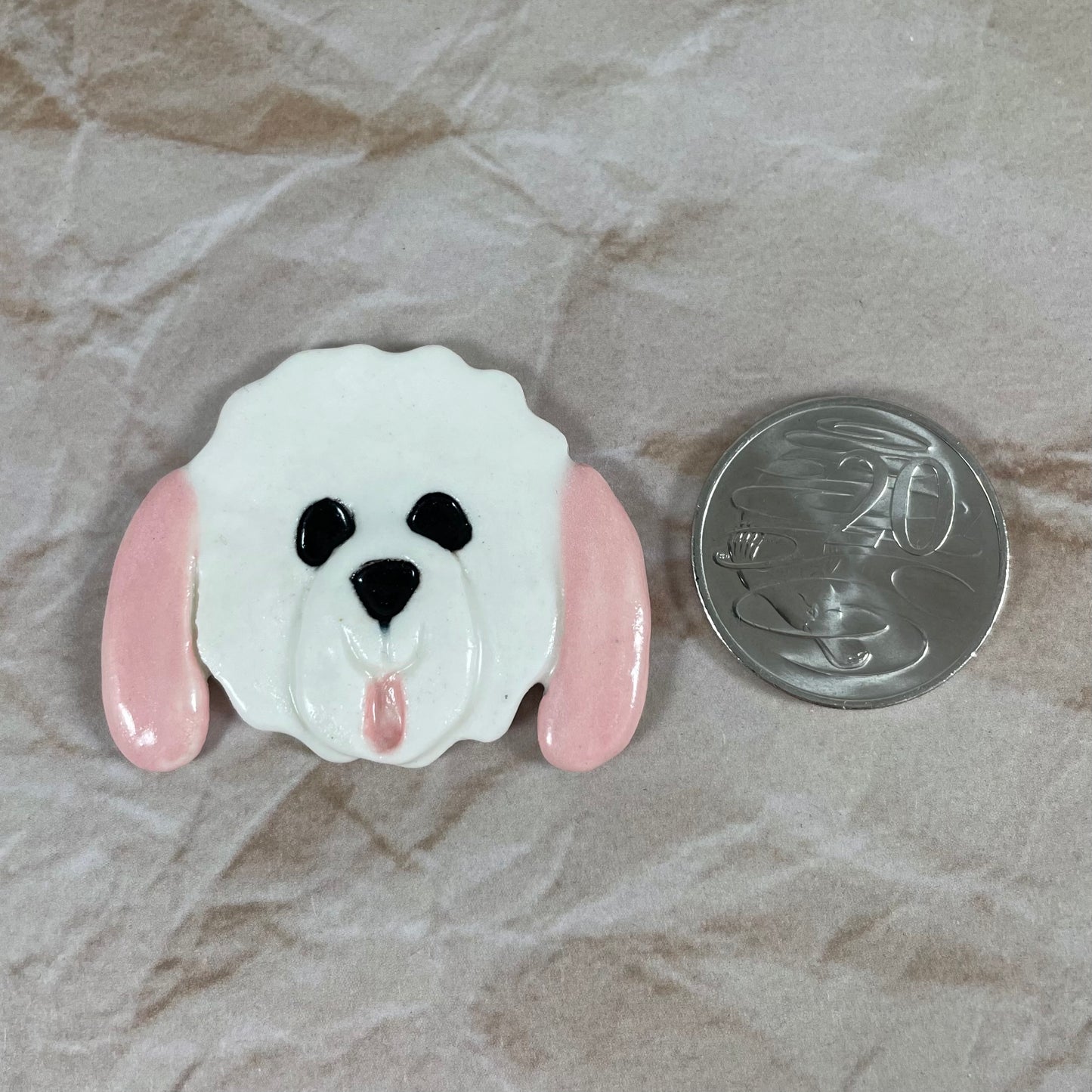 Dog's head in glossy white porcelain with long pale pink ears and pink tongue sticking out. Shiny black eyes and nose. Muzzle area is slightly raised to stand out. Photographed next to 20 cent coin, which is smaller than brooch.