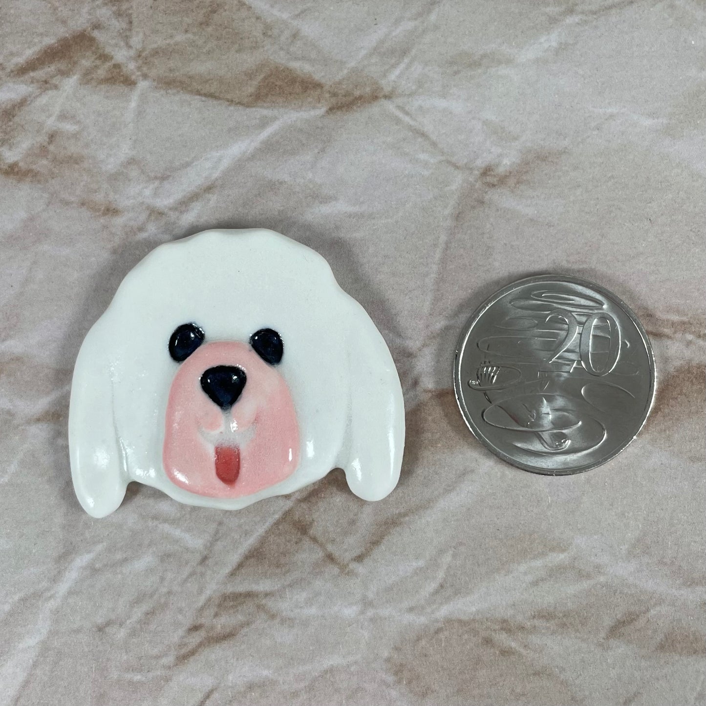 Dog's head in glossy white porcelain with long ears and pink tongue sticking out. Shiny black eyes and nose. Muzzle area is pale pink and slightly raised to stand out. Photographed next to 20 cent coin, which is smaller than brooch.