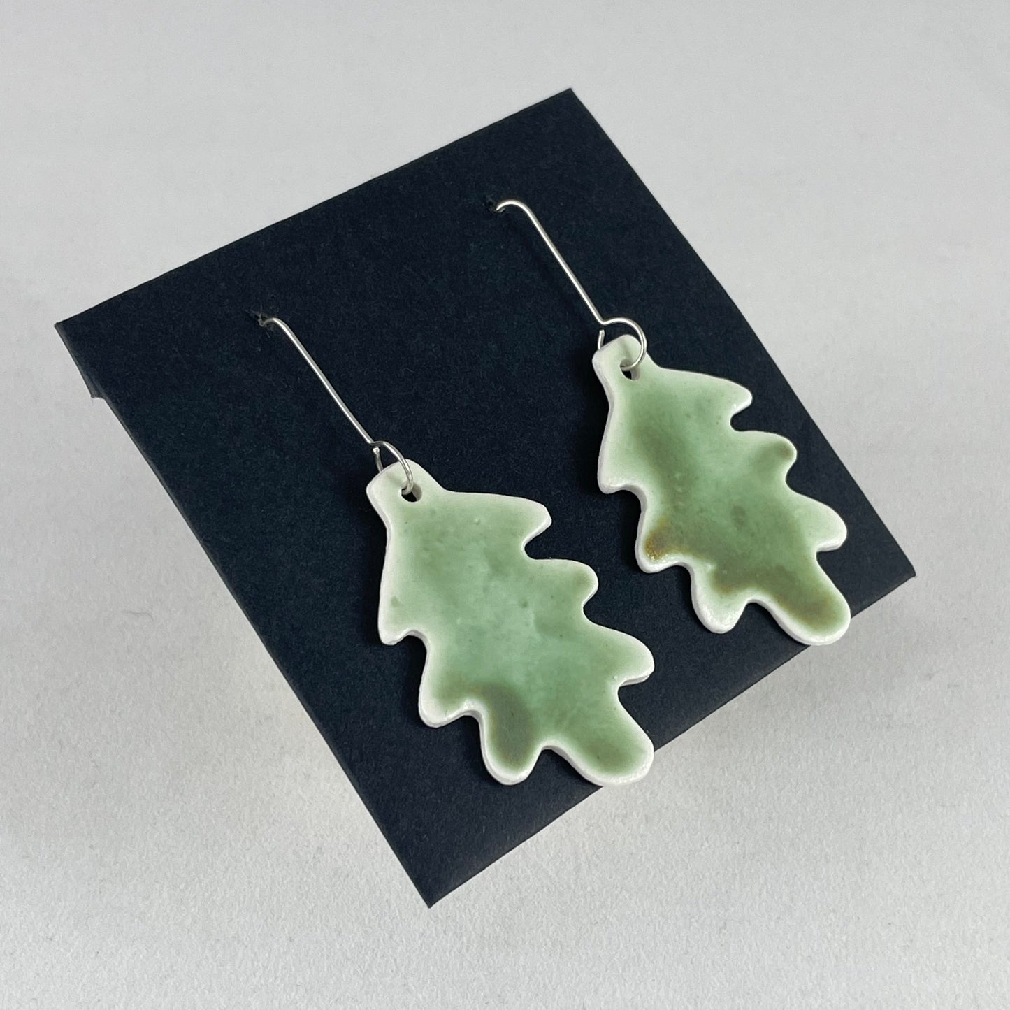 A pair of handmade porcelain earrings in oak leaf shape and warm green glaze. Photographed on a black background. On sterling silver earrings hooks. Side view