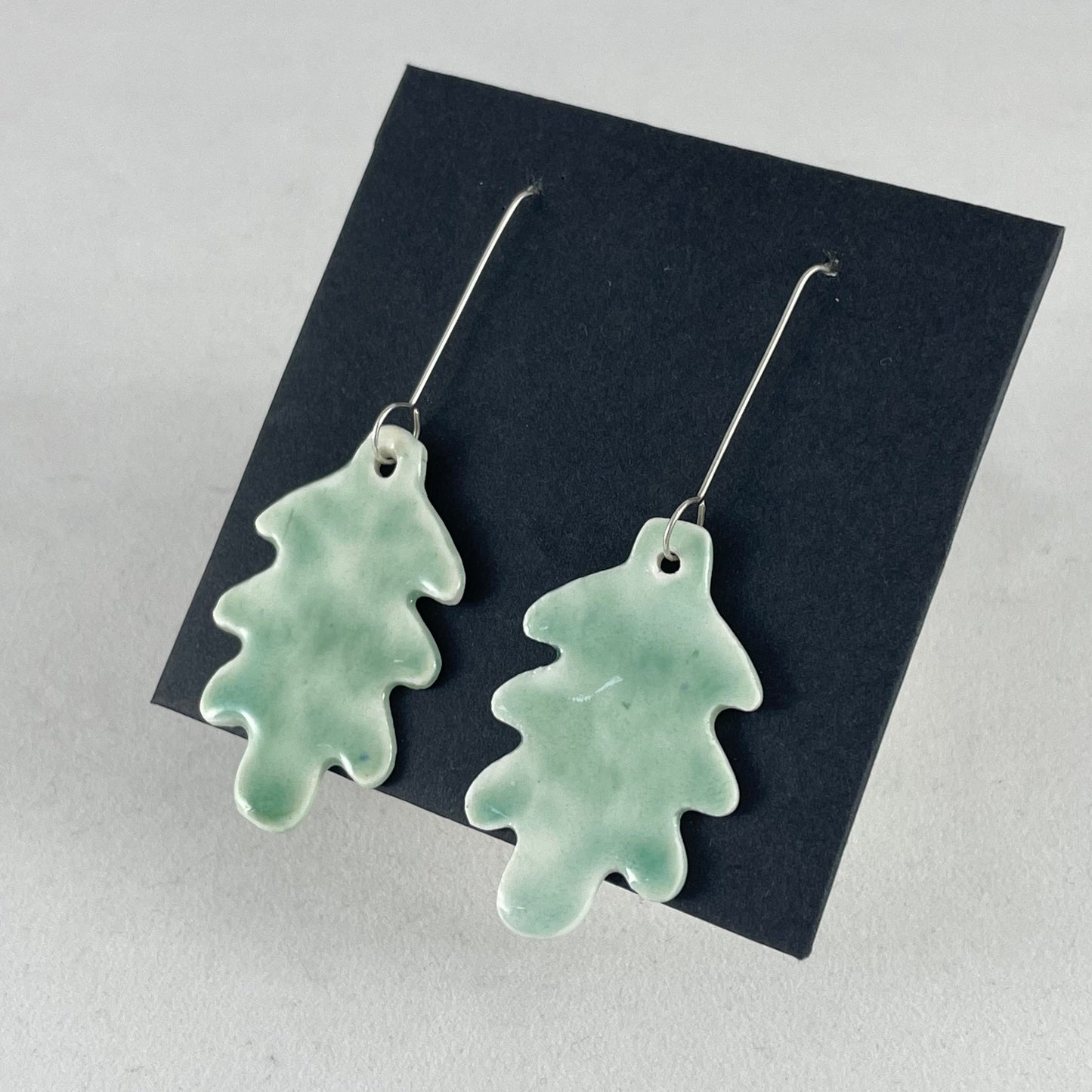 A pair of handmade porcelain earrings in oak leaf shape and glossy mint green glaze. Photographed on a black background. On sterling silver earrings hooks. Side view