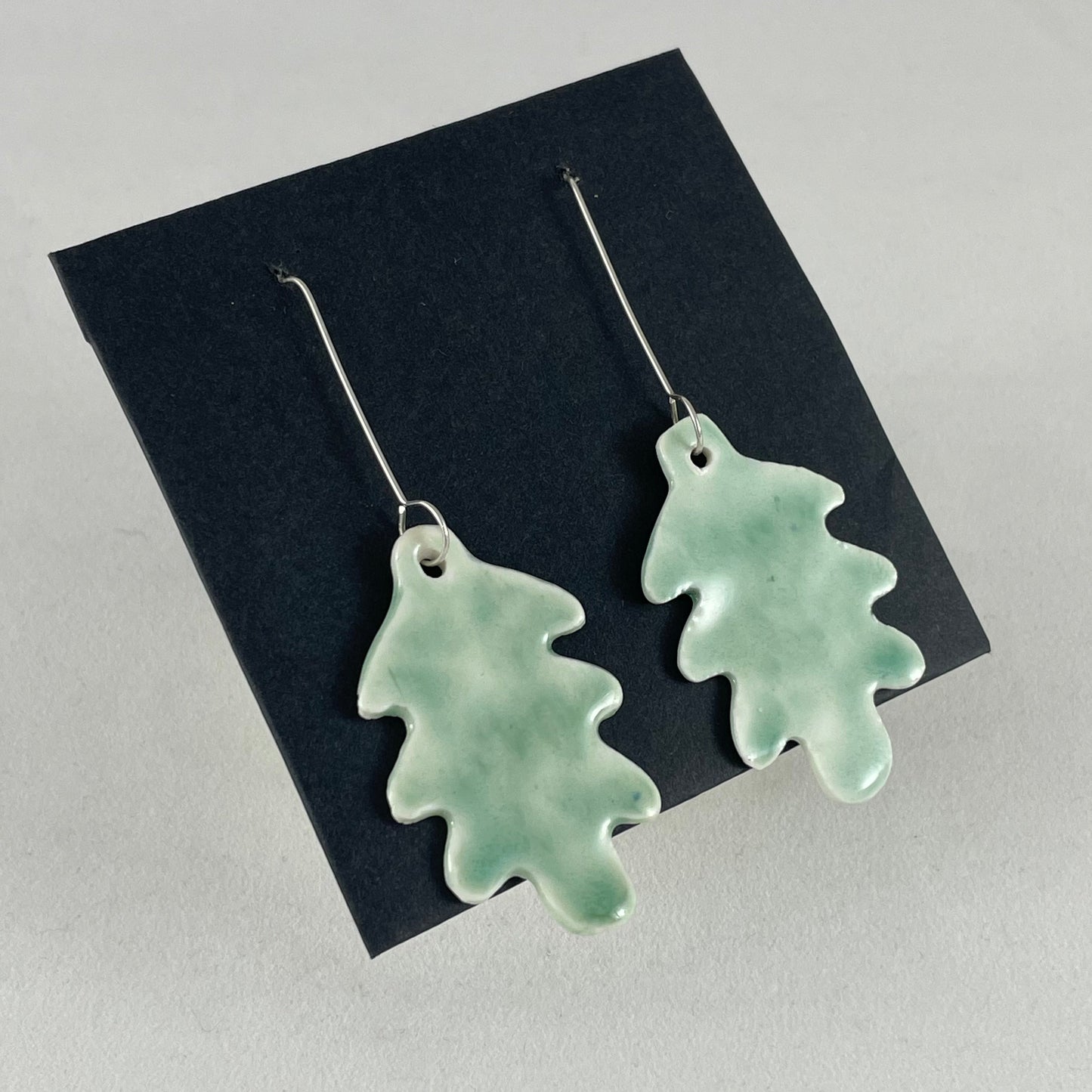 A pair of handmade porcelain earrings in oak leaf shape and glossy mint green glaze. Photographed on a black background. On sterling silver earrings hooks. Side view