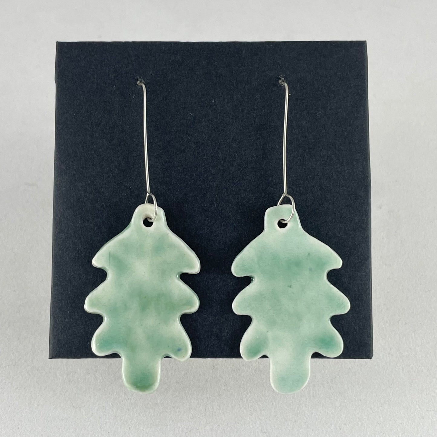 A pair of handmade porcelain earrings in oak leaf shape and glossy mint green glaze. Photographed on a black background. On sterling silver earrings hooks.