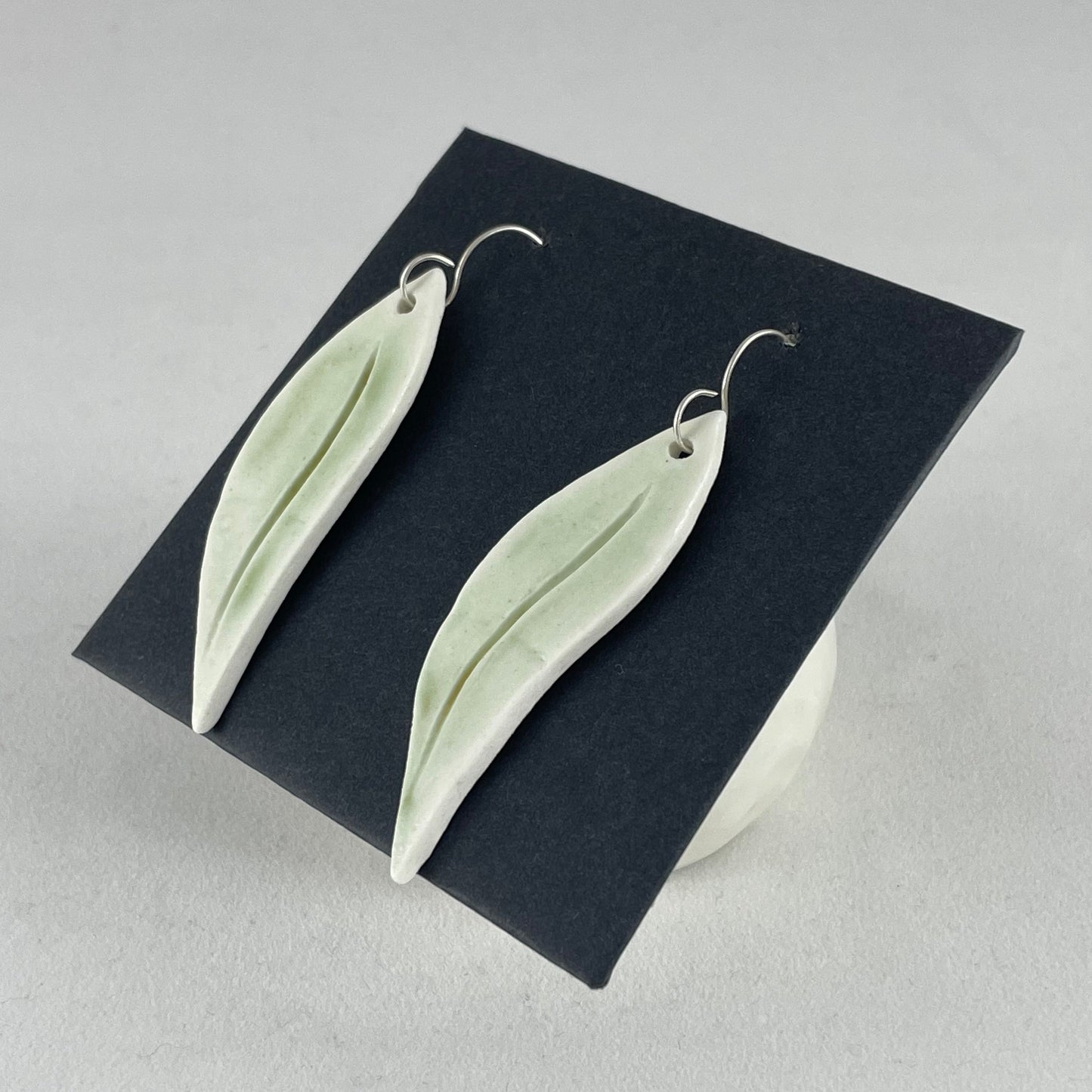 A pair of hand formed porcelain earrings in organic gum leaf shape. Photographed on a black background. On sterling silver earrings hooks. Side view
