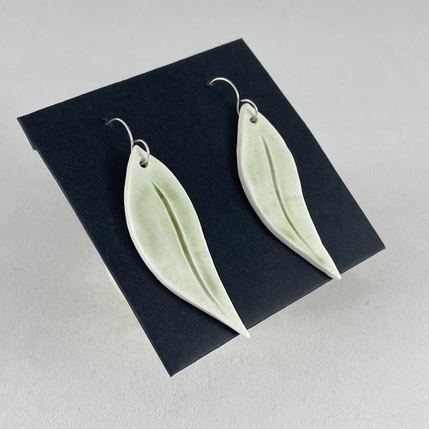 A pair of hand formed porcelain earrings in organic gum leaf shape. Photographed on a black background. On sterling silver earrings hooks. Side view