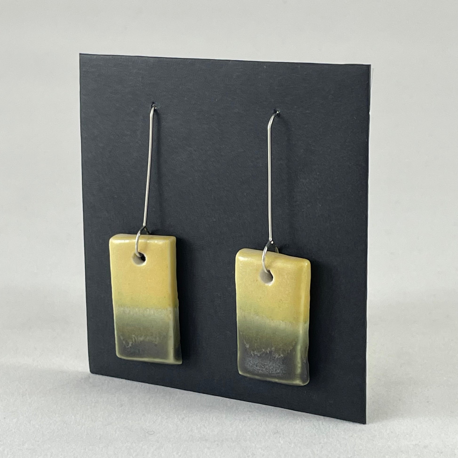 Pair of hand formed porcelain pendant earrings. Yellow and warm green bands of colour on rectangle shape. Sterling silver hooks. Side view