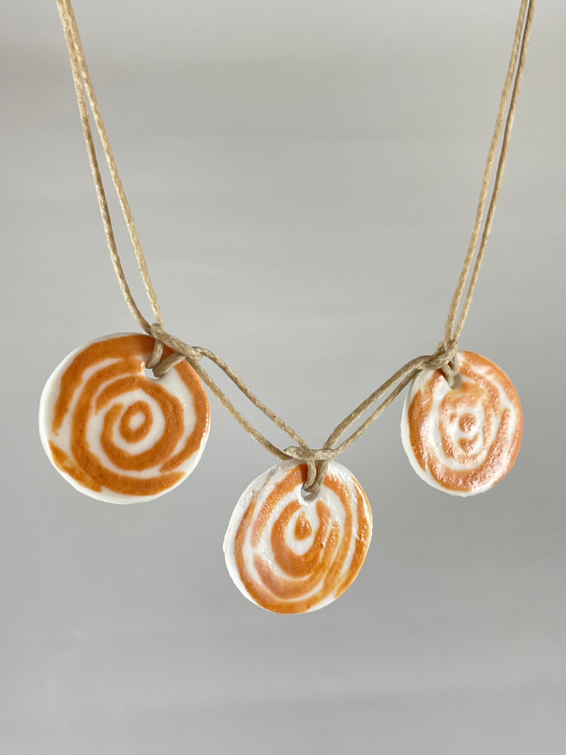 Necklace with three circular porcelain pendants on natural coloured thread, hanging in front of a white background. Orange rose pattern painted on white porcelain. Photographed from left side to show thin profile of porcelain discs