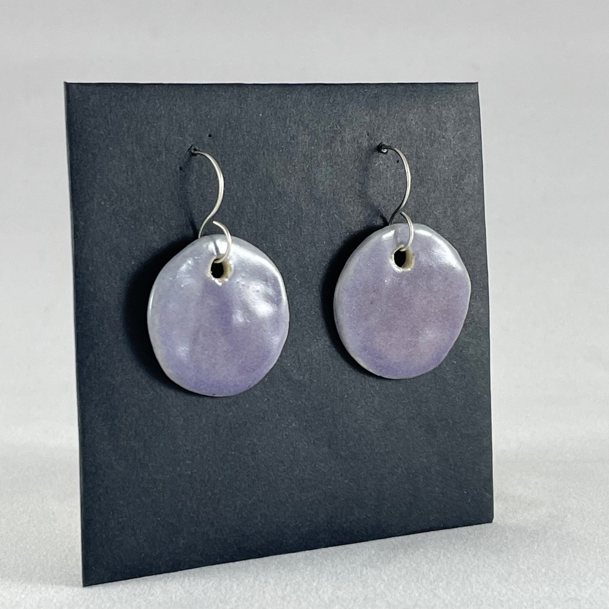 Side view of a pair of circular shaped porcelain earrings in purple. On sterling silver earrings hooks.