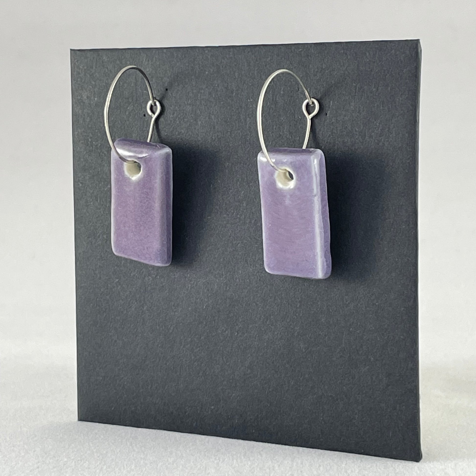 Side view of a pair of rectangular shaped porcelain hoop earrings in purple. On sterling silver earrings hooks.