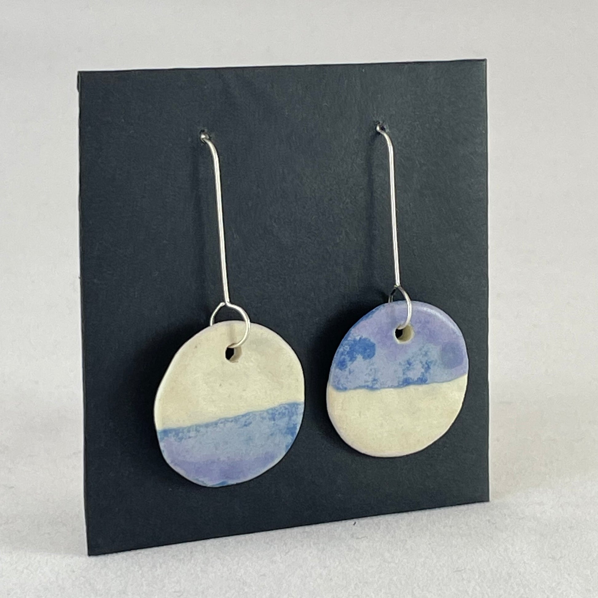 Side view of a pair of circular shaped porcelain earrings in blue and white. On sterling silver earrings hooks.