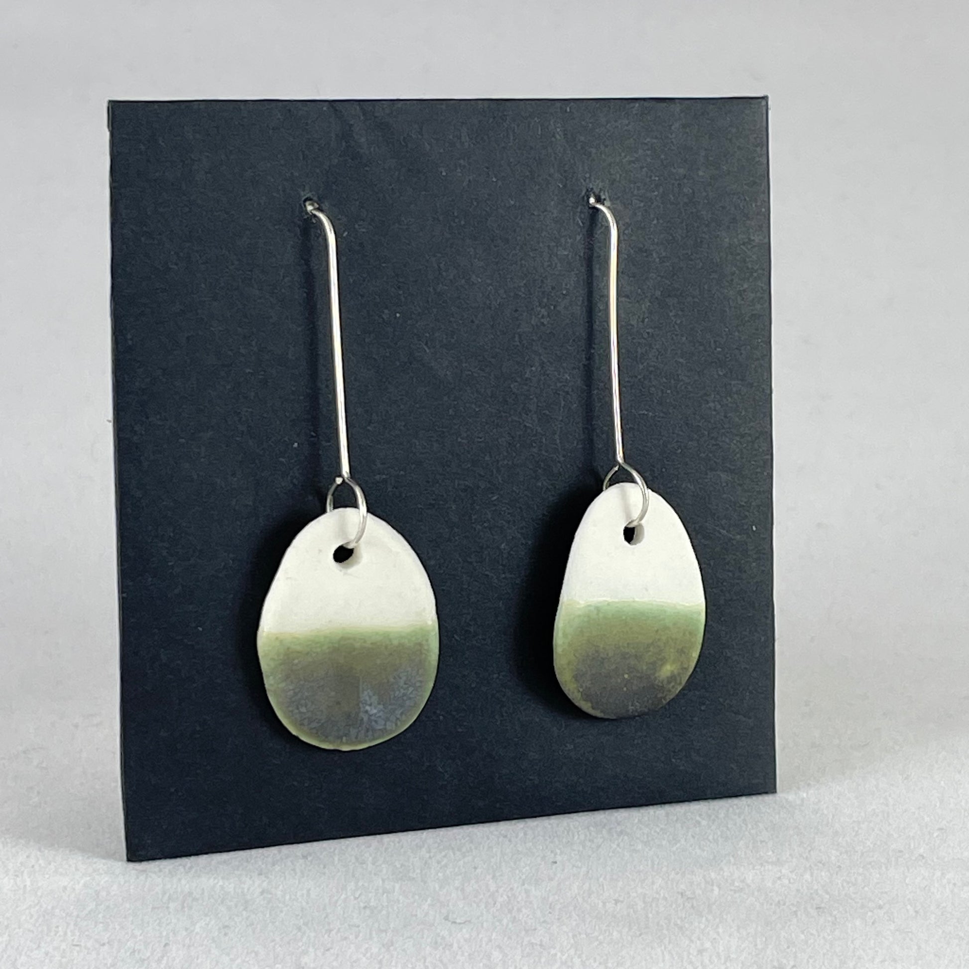Side view of a pair of oval shaped porcelain earrings in dark green and white. On sterling silver earrings hooks.